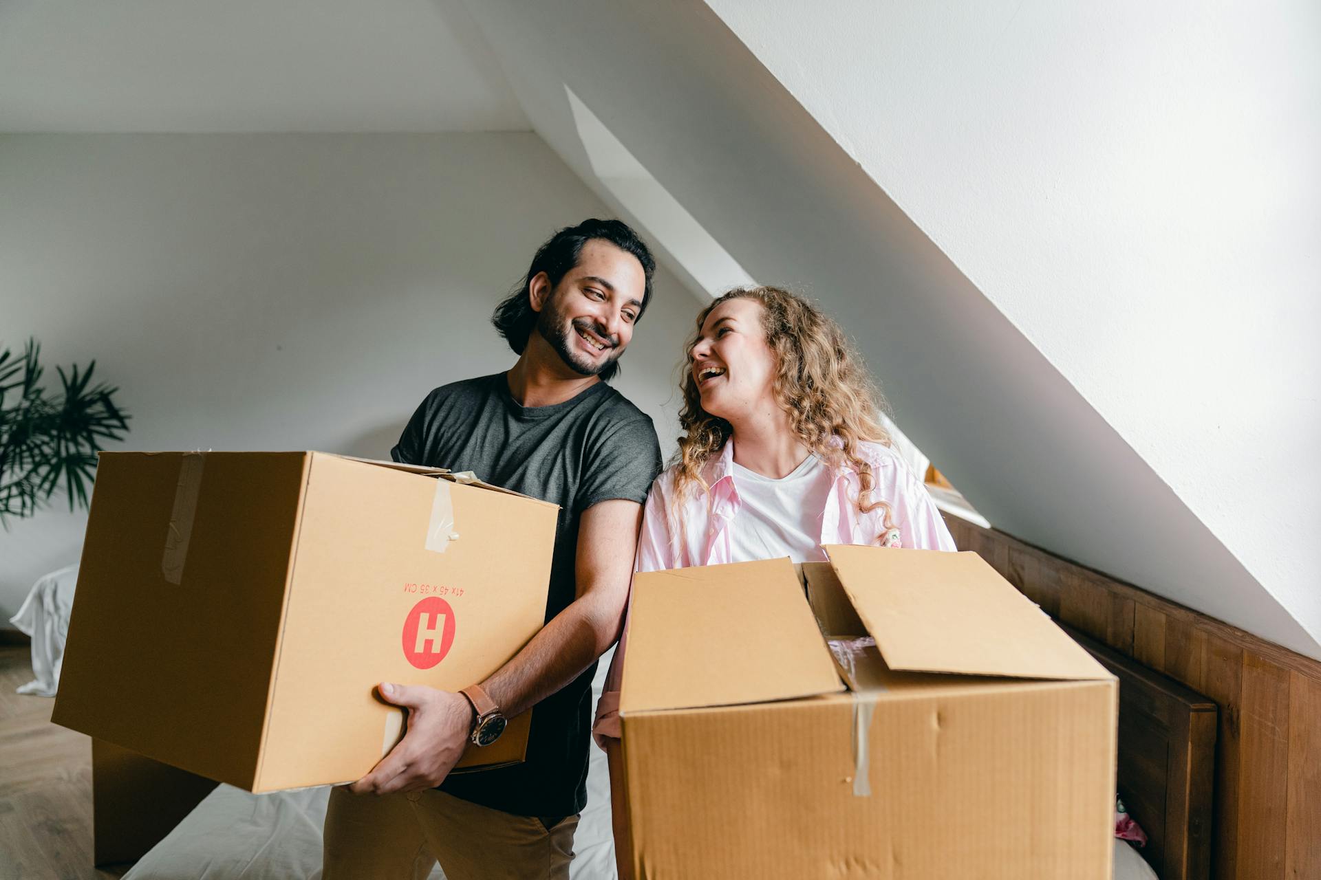  A smiling couple, a man with dark hair and a woman with curly blonde hair, stands inside a bright room, likely an attic or a room with sloped ceilings, holding cardboard moving boxes. They are looking happily at each other.