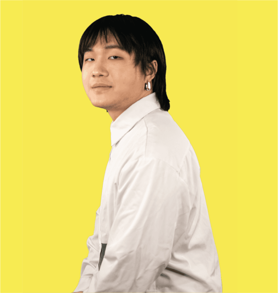 A person with dark hair styled in a side-swept manner wears a white long-sleeved shirt against a solid yellow background. The individual is positioned in profile, facing left, with their gaze directed forward. They are wearing small, silver hoop earrings. The shirt has a collar and appears to be made of a smooth, light-colored fabric. No other objects or text are visible in the image.