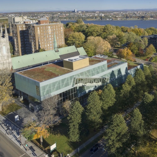 Modern glass building with a green roof, surrounded by trees, a church, and other buildings, near a large body of water.