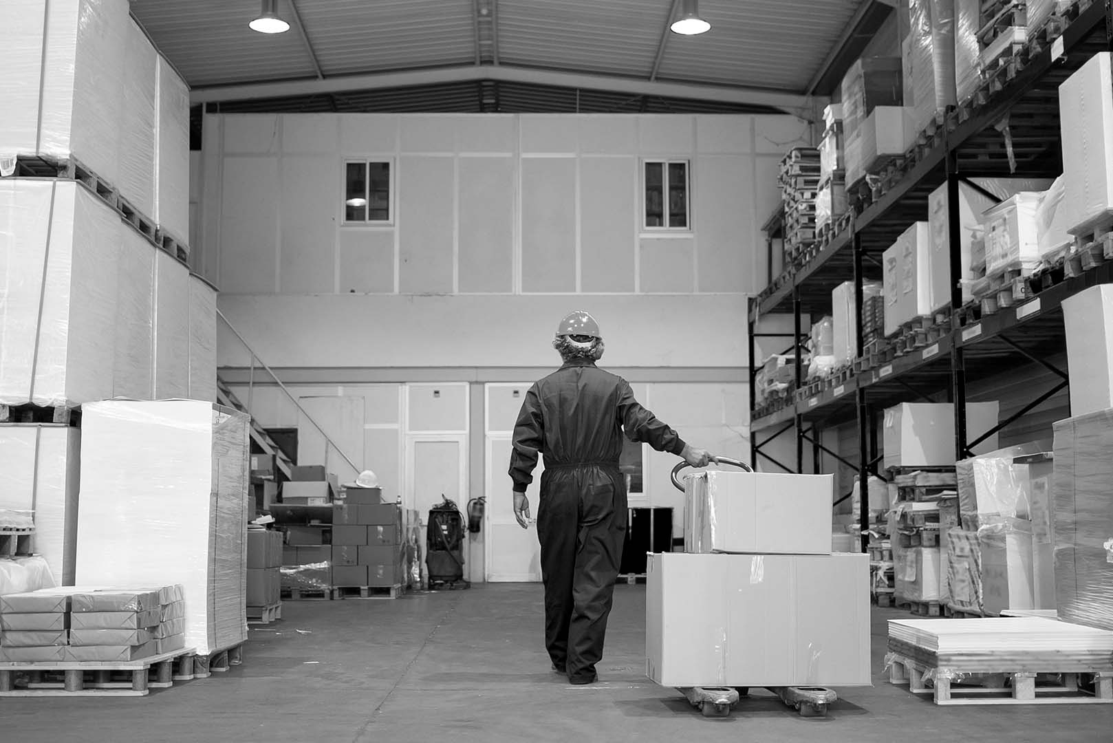 A man wheeling a pallet loaded with boxes through a warehouse. The background shows tall shelves filled with goods, suggesting an active industrial or storage environment.