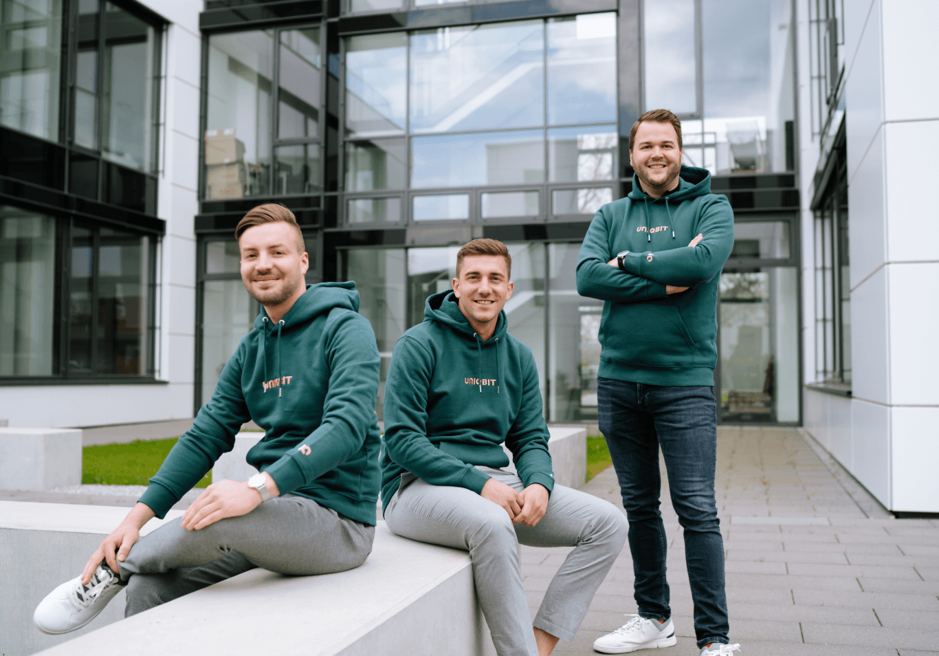 Zwei männer sitzen auf einem Steinblock vor einem modernen Gebäude die dritte Person steht daneben