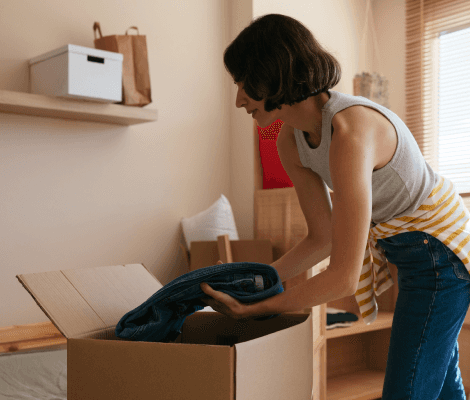 A woman with short hair is placing a folded pair of jeans into a cardboard box. She is wearing a sleeveless top and has a striped apron tied around her waist. The room is well-lit with natural light, and there are shelves and a few bags in the background, indicating a tidy living space.