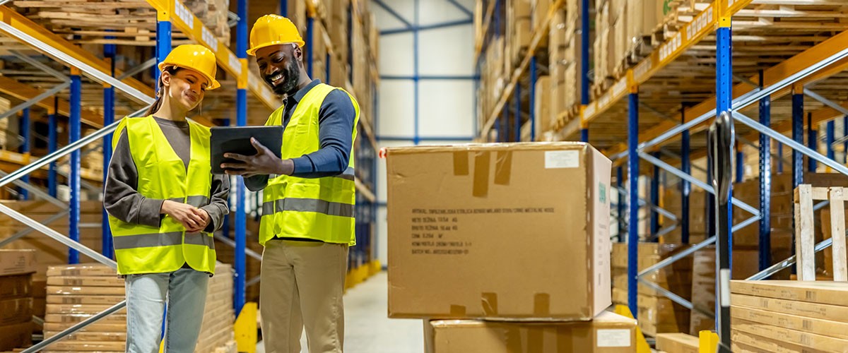 Two workers in safety gear inspect inventory in a warehouse aisle surrounded by boxes and shelves.