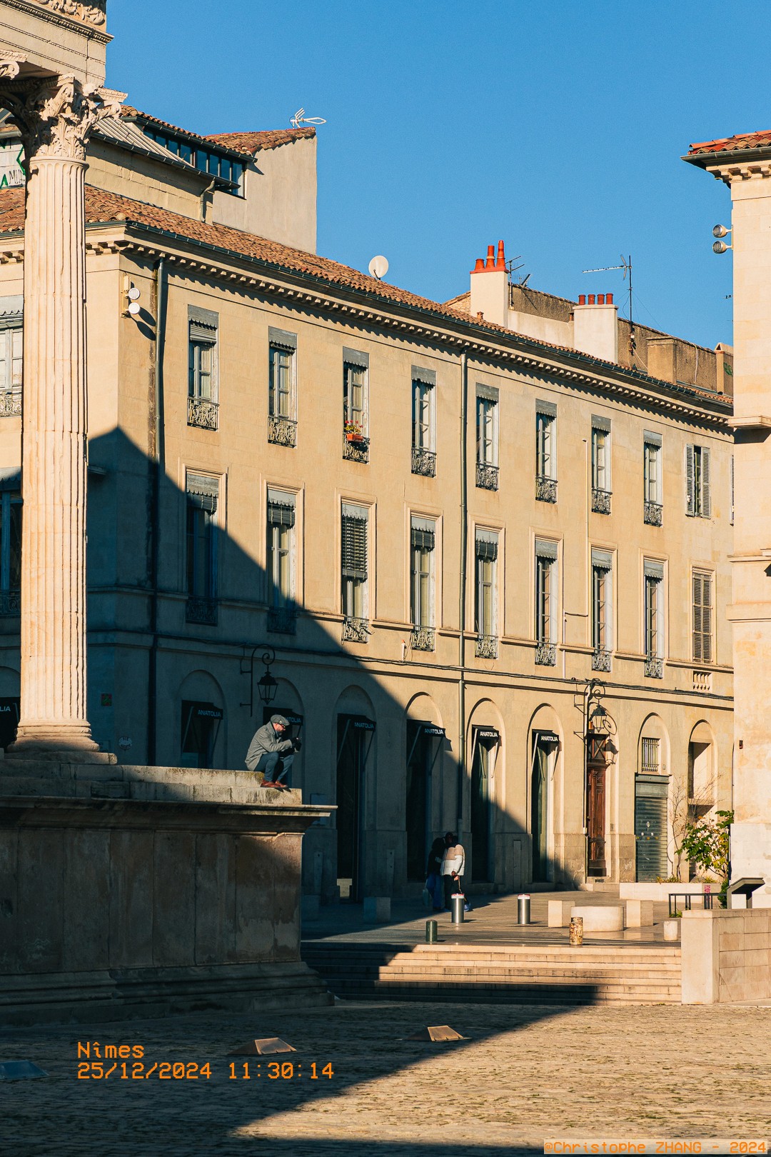Un homme assis sur un muret en pierre de la Maison Carrée, lisant dans la lumière du matin à Nîmes.