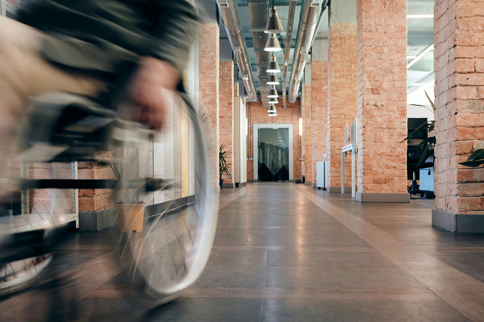 A modern office corridor with exposed brick columns and industrial-style pendant lighting. A blurred figure of a wheelchair user moves past glass-walled offices on the left, conveying movement and workplace activity. The space features polished concrete floors, exposed ductwork on the ceiling, and natural light streaming through windows at the end of the hallway, creating a contemporary industrial aesthetic.