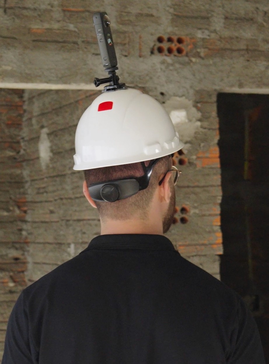 Vista traseira de um colaborador usando um capacete de segurança branco com uma câmera de Captura da Realidade 360° acoplada no topo. Ele está em um ambiente de obra com paredes de tijolos aparentes, demonstrando como o registro é feito de forma rápida e com as mãos livres.