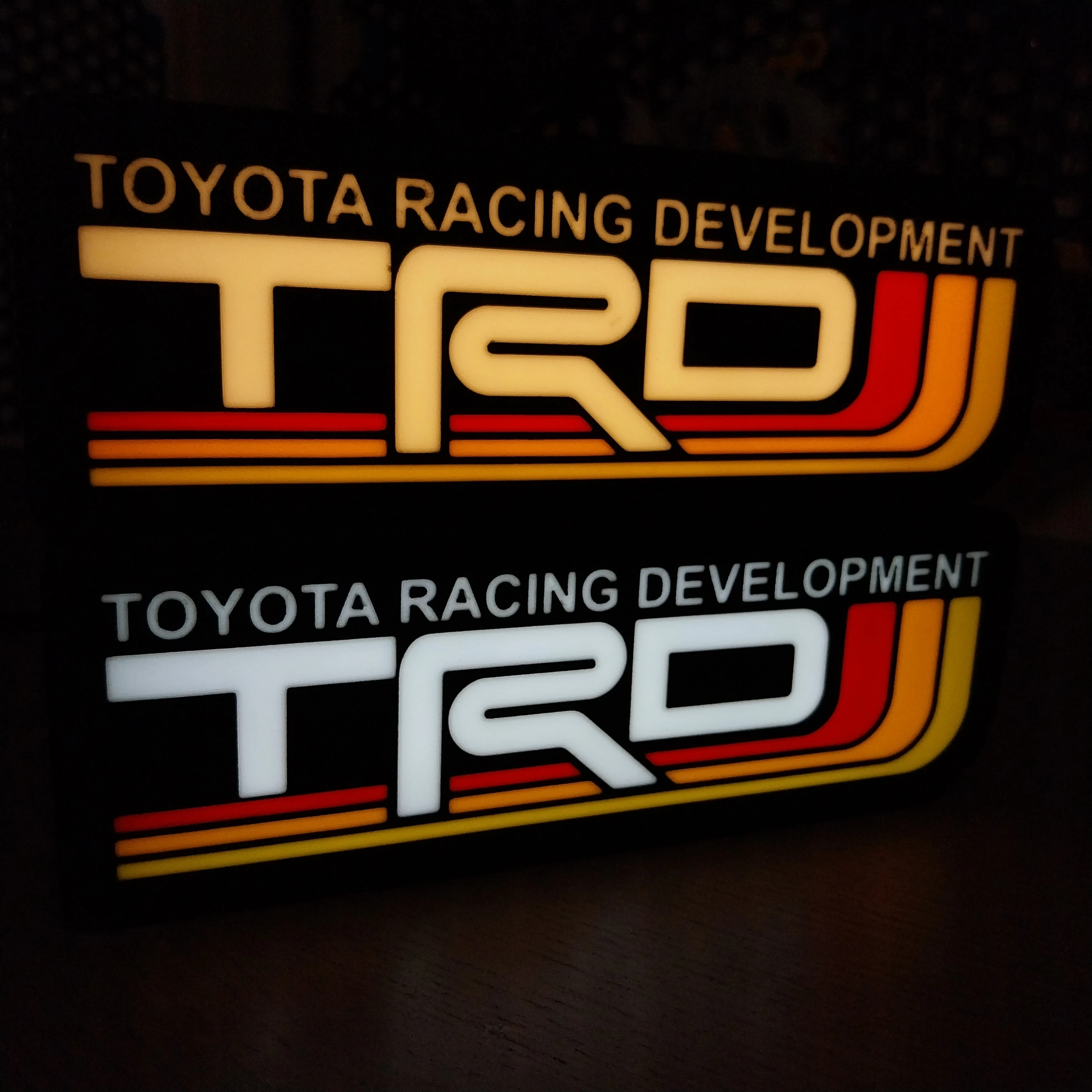 Two TRD (Toyota Racing Development) stickers placed over a magazine featuring car suspension, engine components, and a red Toyota sports car. Emphasizes TRD’s connection to performance tuning and custom parts.