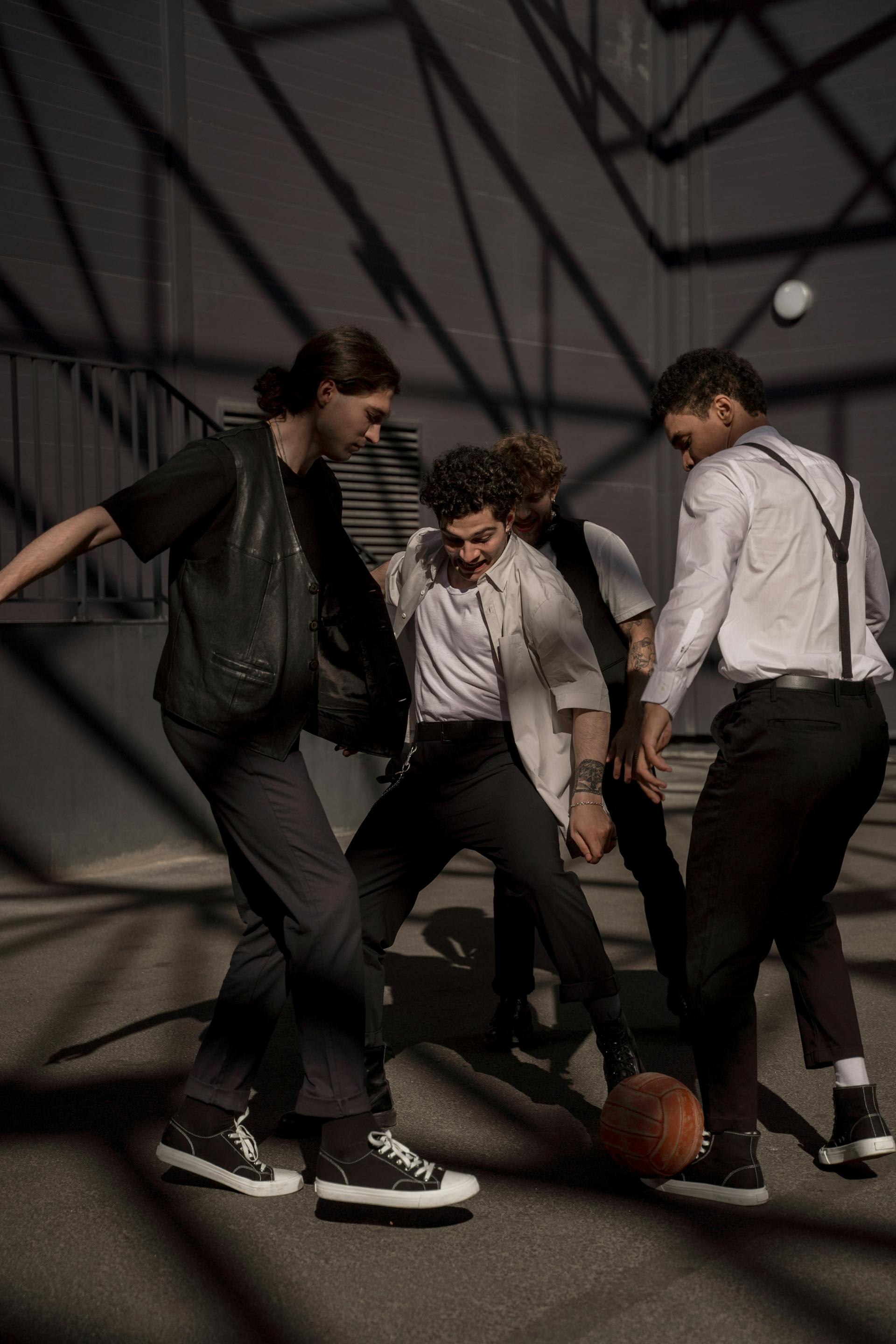 Four men in a dynamic action shot, appearing to play or compete over a vintage leather ball on an asphalt ground with dramatic geometric shadows.