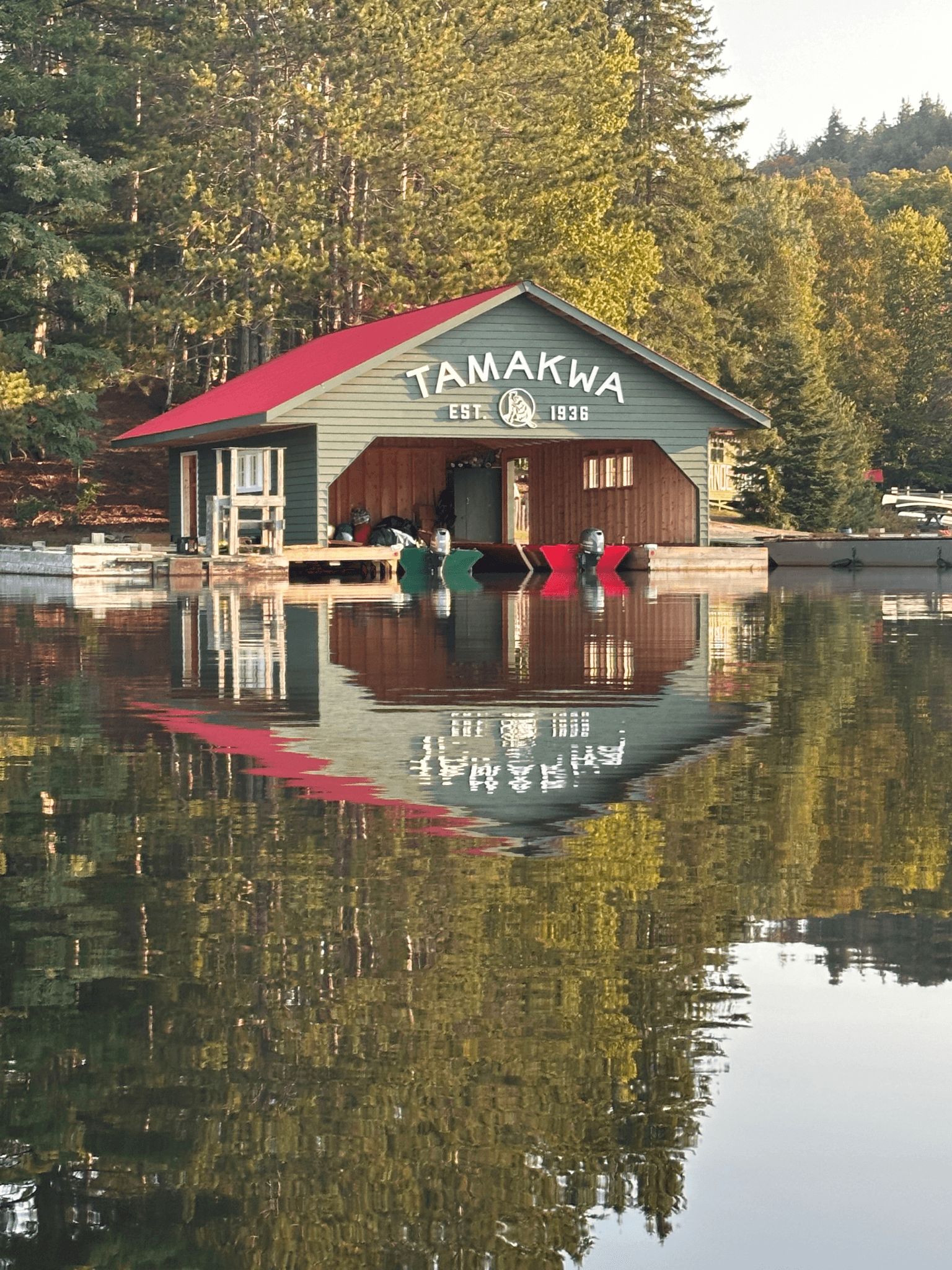 Tamakwa boathouse on a calm lake, reflecting in water, surrounded by Algonquin Park forest.