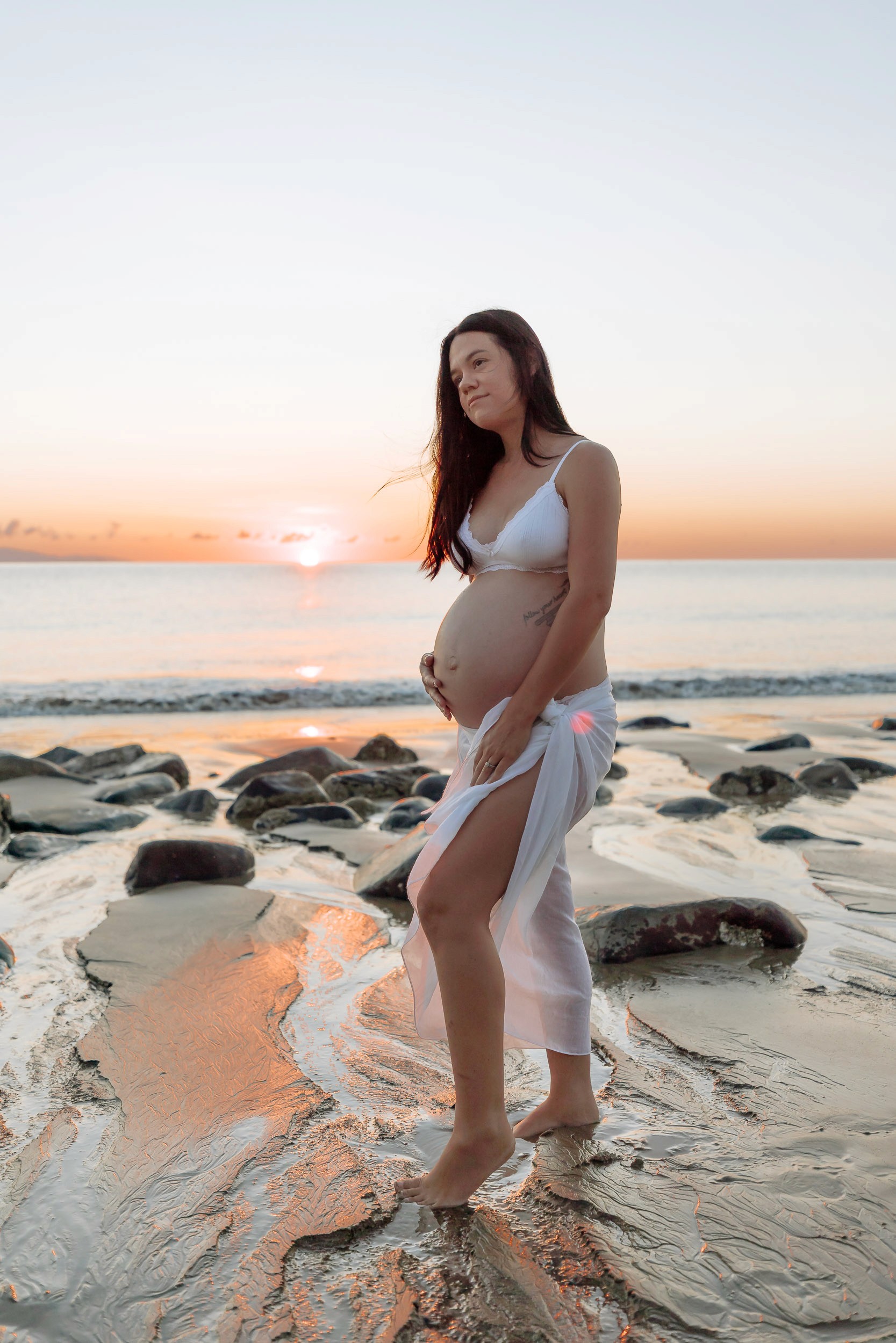 Outdoor solo maternity session on a quiet beach at first light, glowing sunrise sky and still water framing an intimate, natural pregnancy moment.