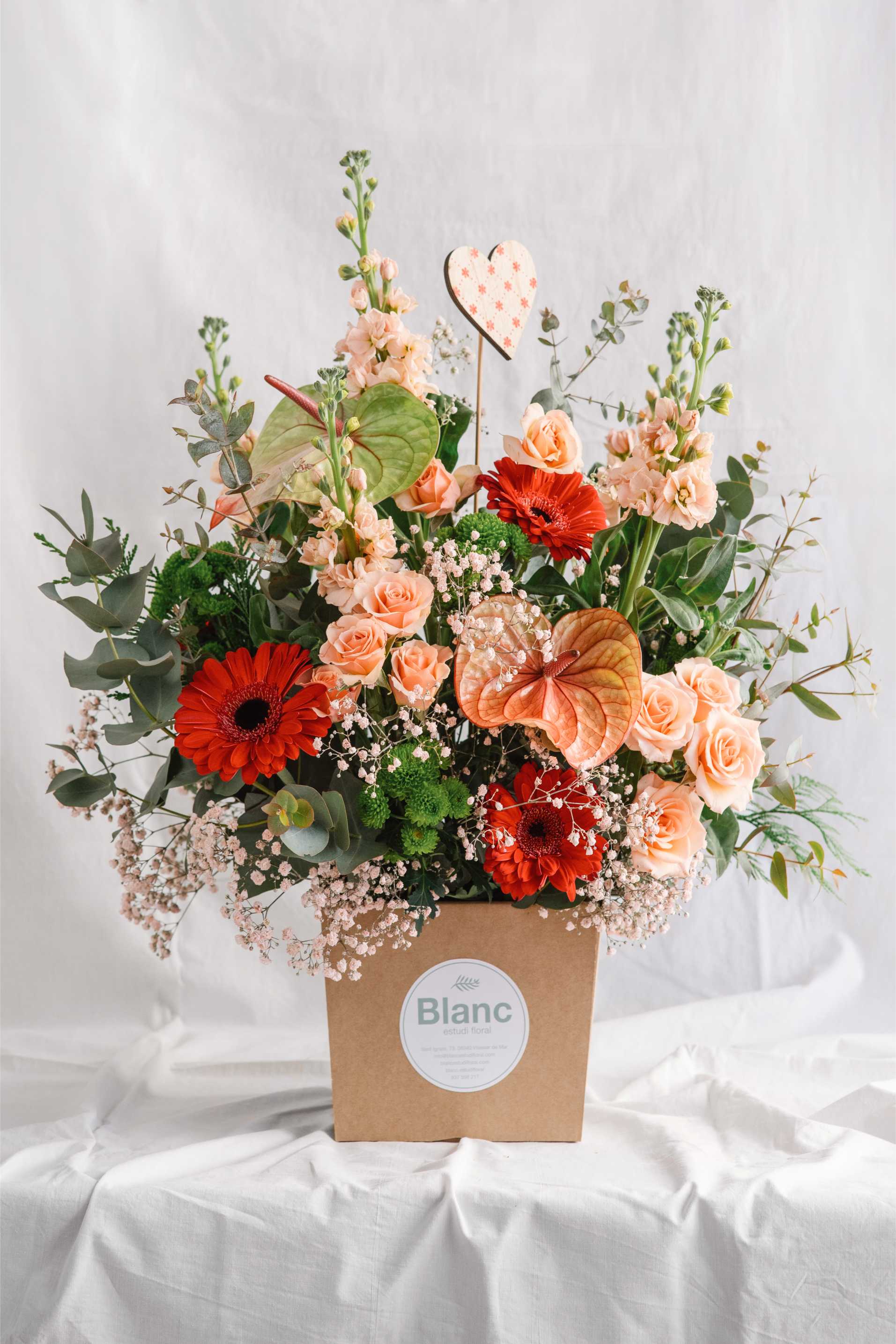 Arranjament floral de gran format amb roses préssec, gerberes vermelles, anthurium i verds variats, presentat en una base de cartró kraft amb etiqueta de Blanc Estudi Floral.