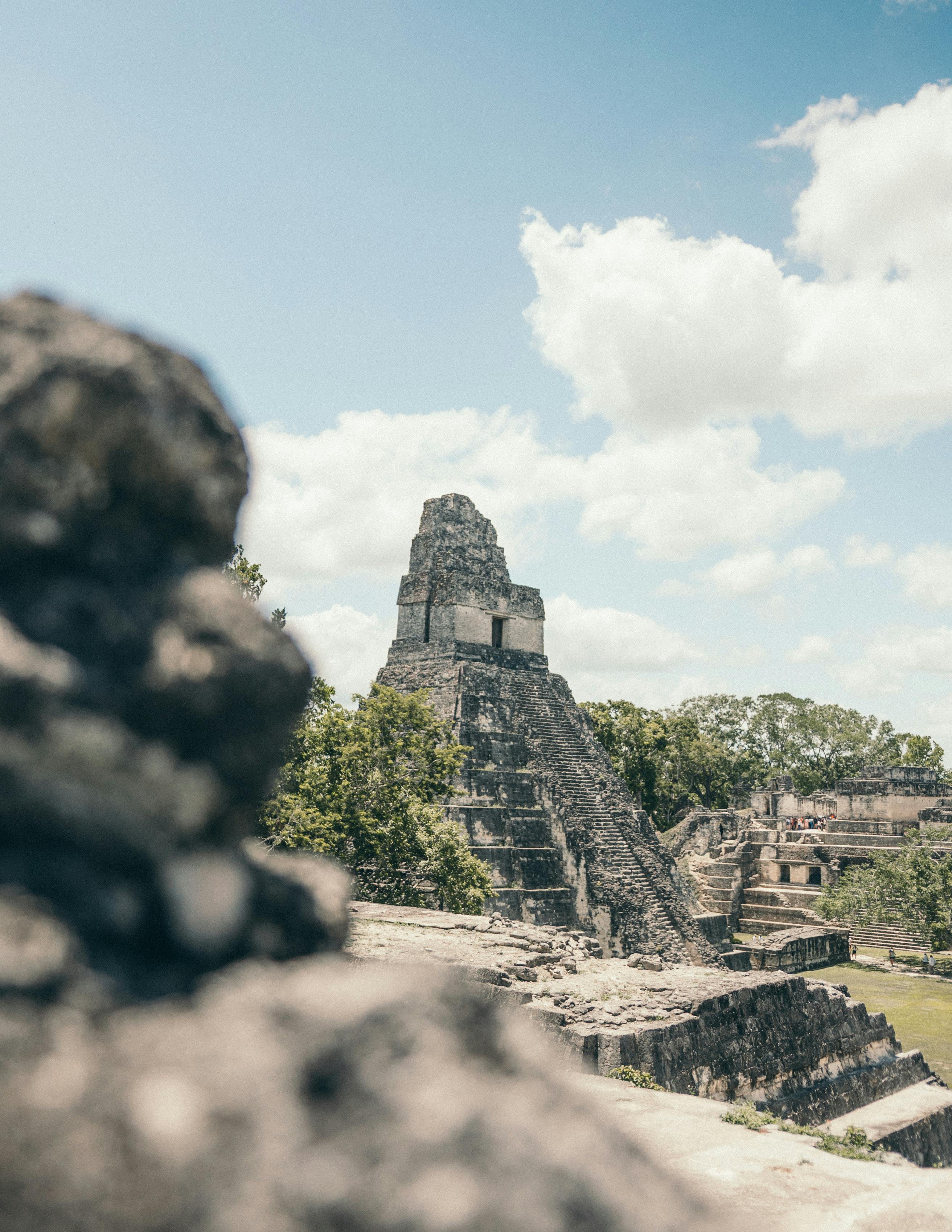 Ancient Mayan pyramid at Tikal rises against a clear blue sky, surrounded by lush greenery and scattered clouds.