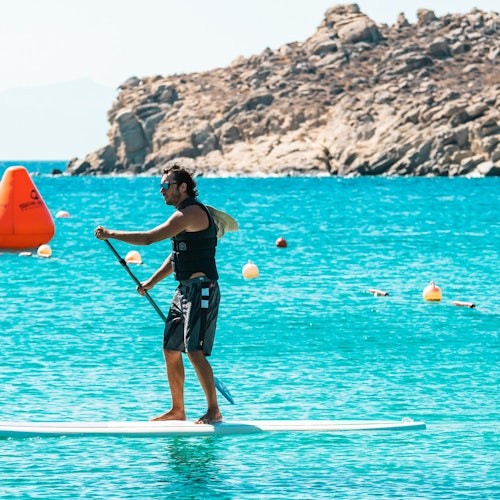 A man paddleboarding on clear blue water near rocky terrain, with buoys in the background.