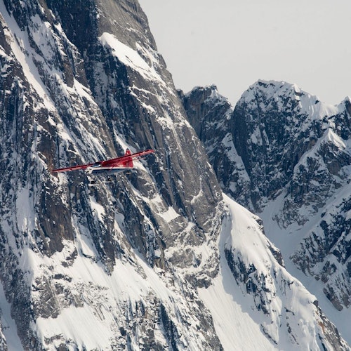 一架紅色飛機在多雲的天空下飛行，靠近雪白、崎嶇的山峰。