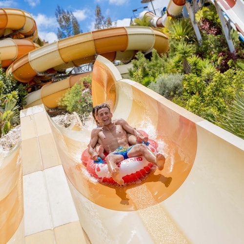 A person riding an inflatable raft down a water slide, with lush greenery and a complex slide structure in the background.