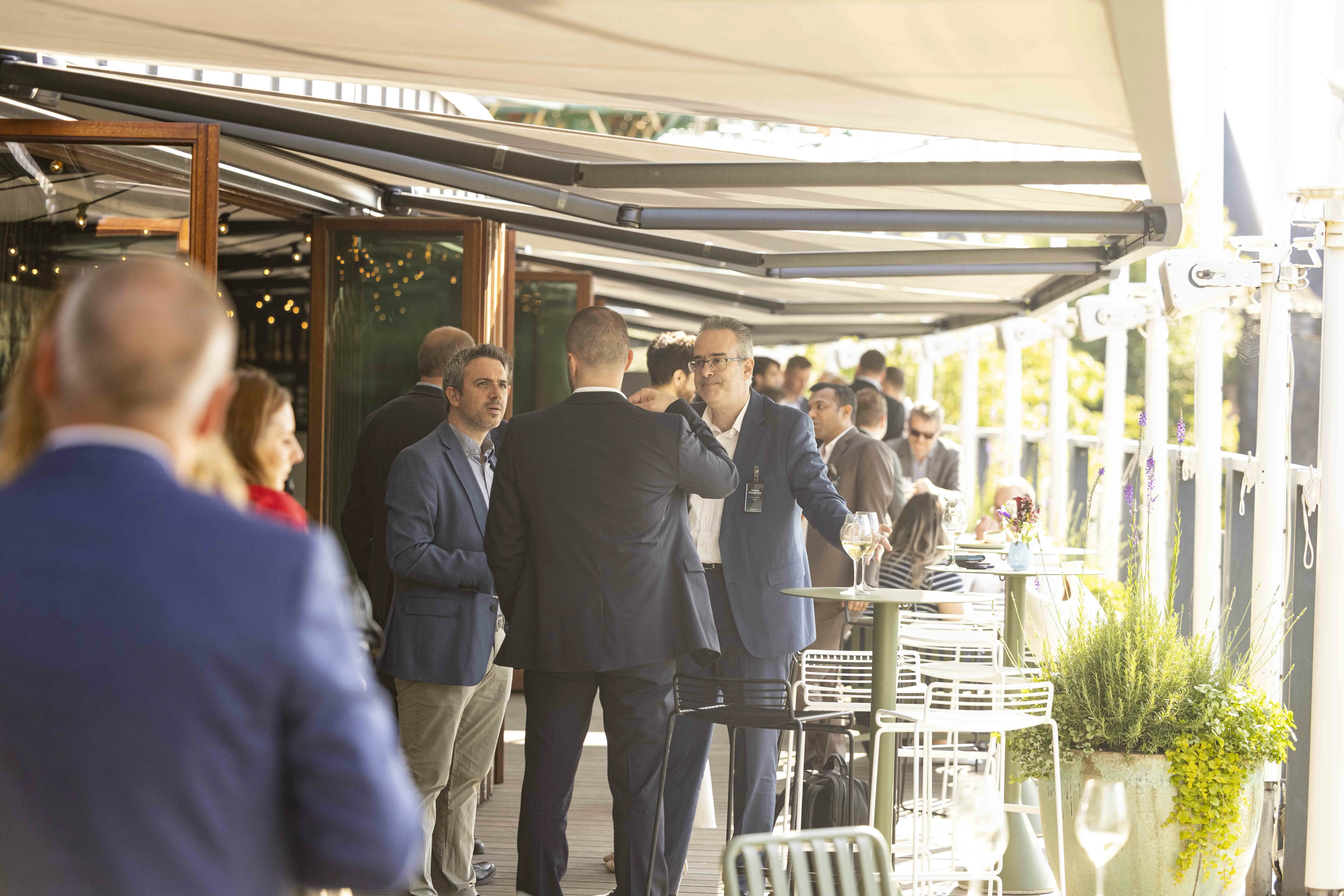 A group of people chatting casually in a modern outdoor setting with greenery and natural light.