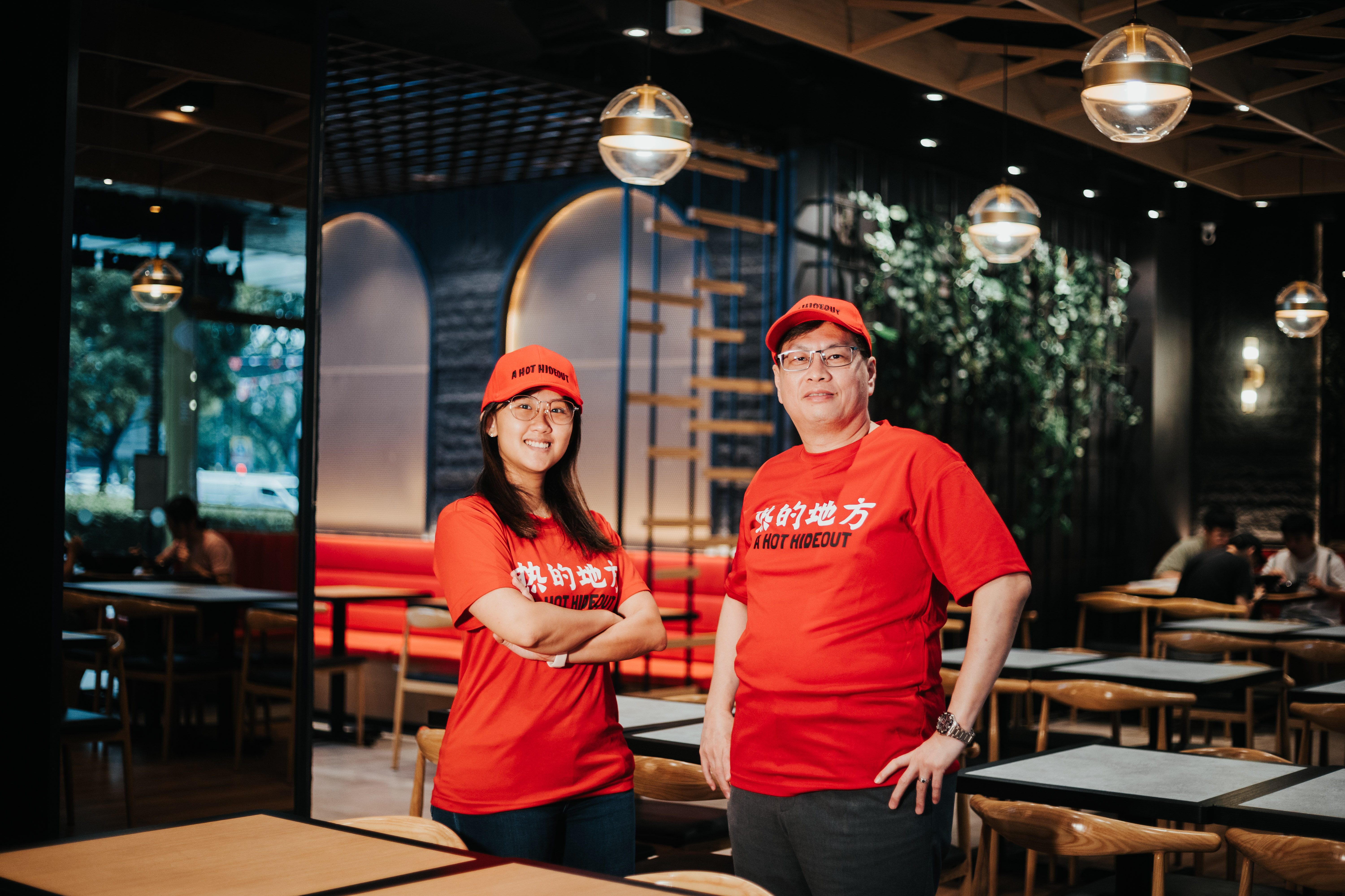 Two smiling employees wearing red shirts and hats stand in a restaurant, with a warm interior and seating behind them.
