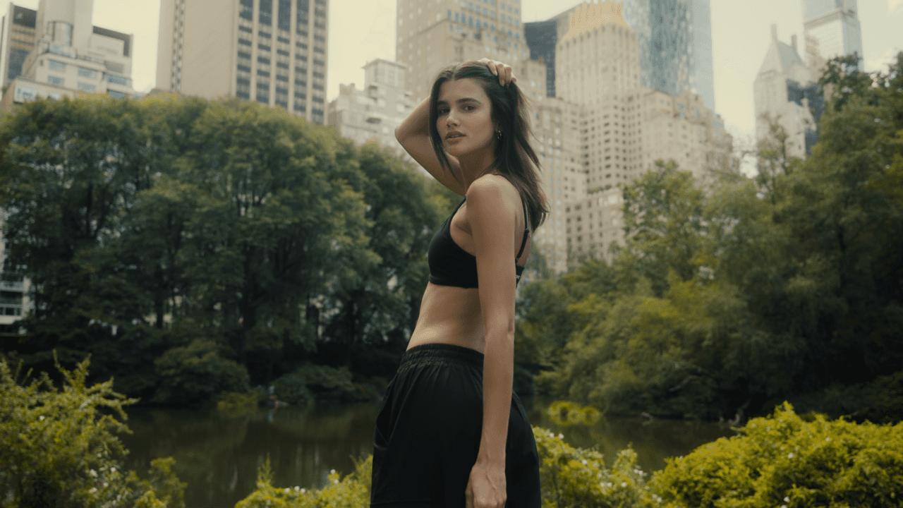 A model standing confidently in a park, with tall buildings and the city's skyline visible behind her