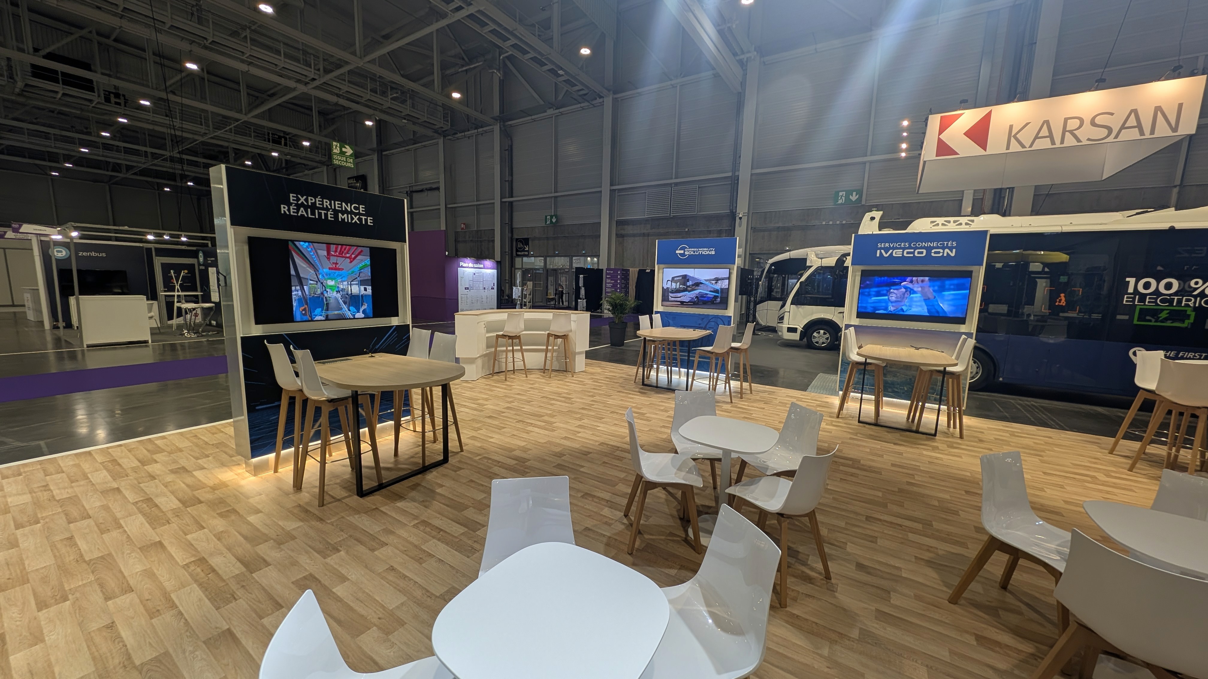 Stand IVECO Bus et Heuliez RNTP 2025 conçu par Expace – vue intérieure espace lounge avec mobilier clair, tables rondes et ambiance identitaire bleue marine