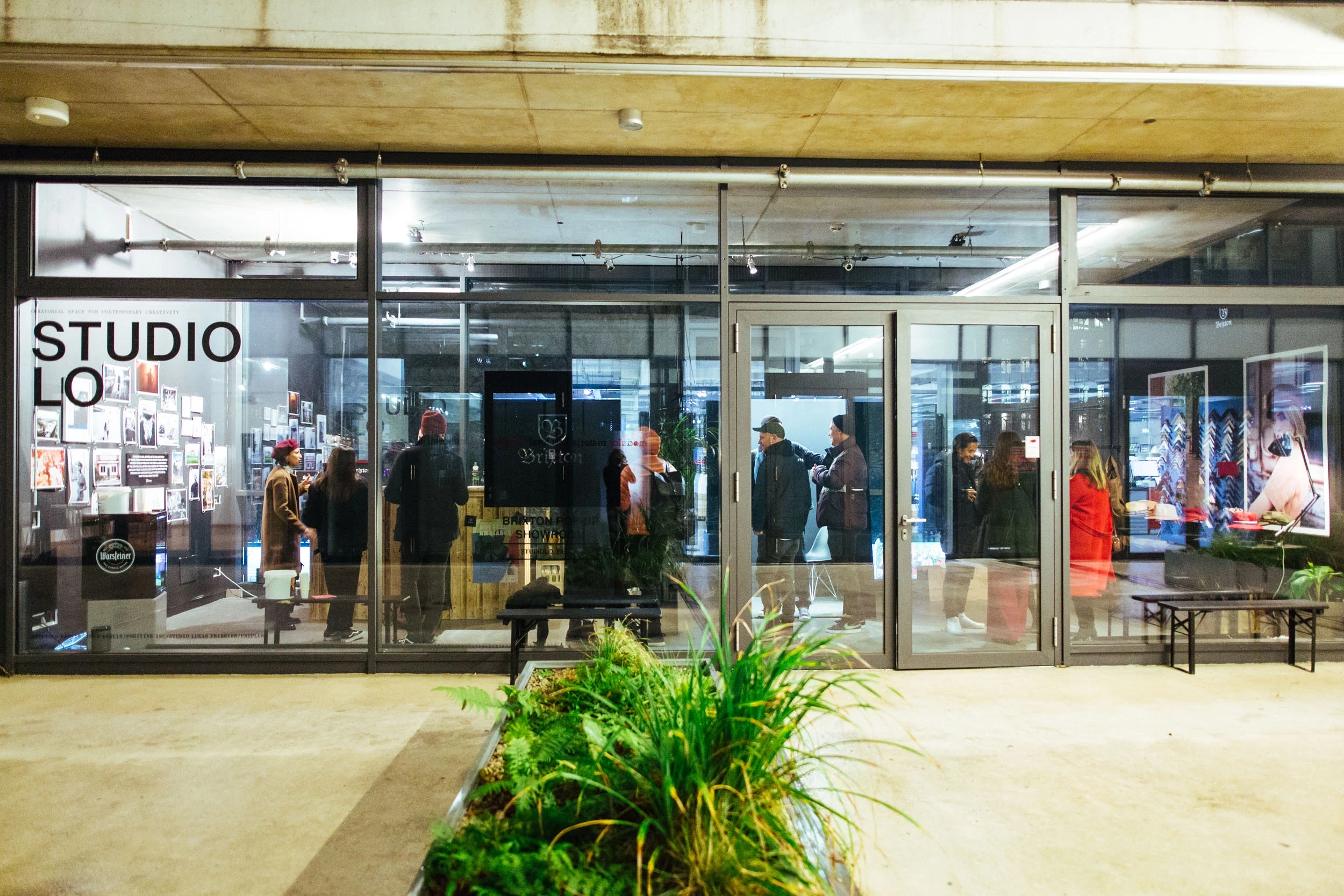 Evening exterior of the BRIXTON pop-up at STUDIOLO with guests visible through the glass facade and a planted bed in front of the entrance.