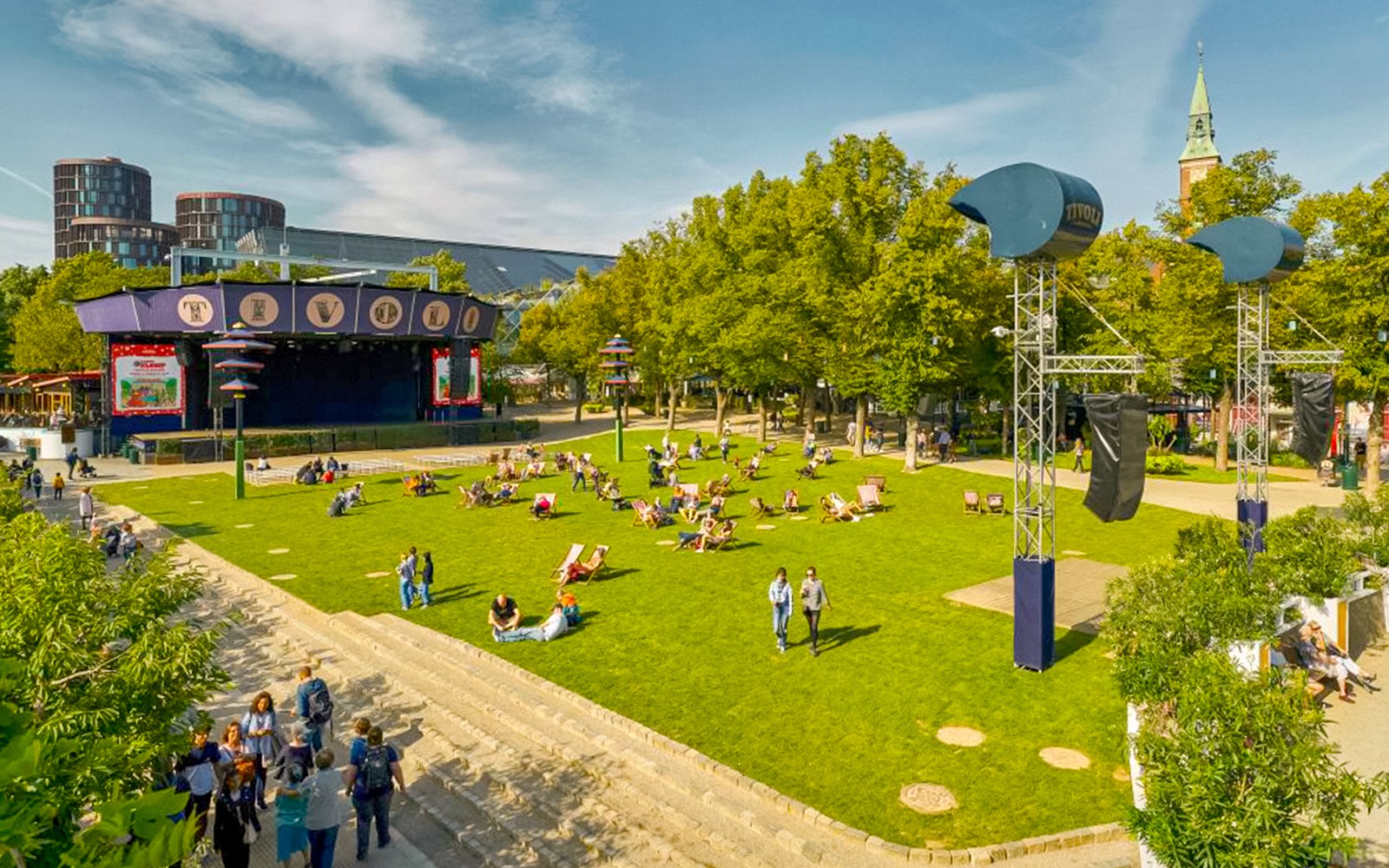 Látogatók pihennek a Tivoli Kertek gyepén, Koppenhágában, a közeli látnivalók és vidámparki játékok között.