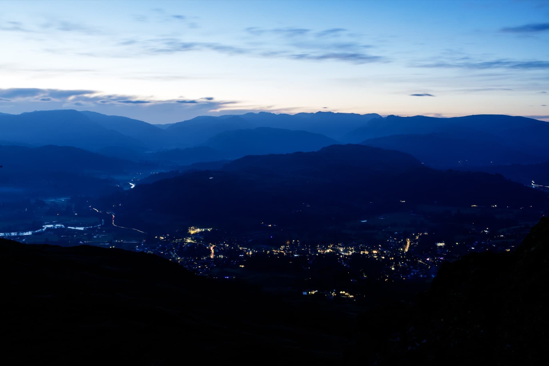 Elevated twilight view from Wansfell Pike showing Lake District settlements illuminated below, layered mountain ridges receding into distance under fading blue sky