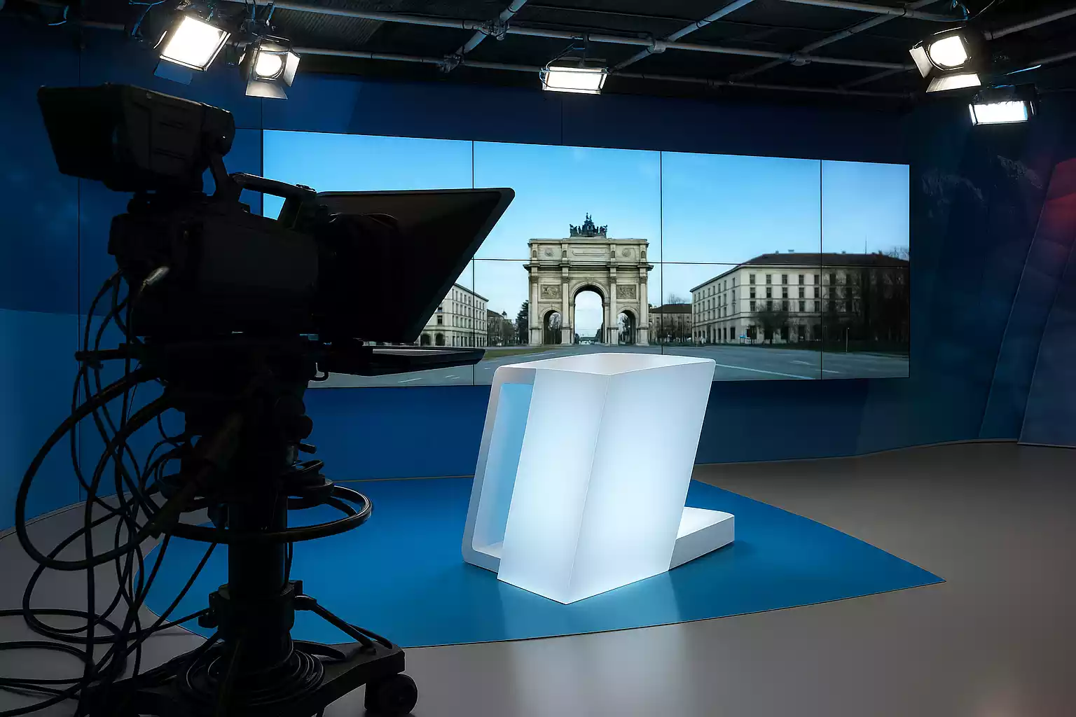 Studio münchen.tv im Studio, hält Moderationskarten vor einem Bildschirm mit Stadtszene