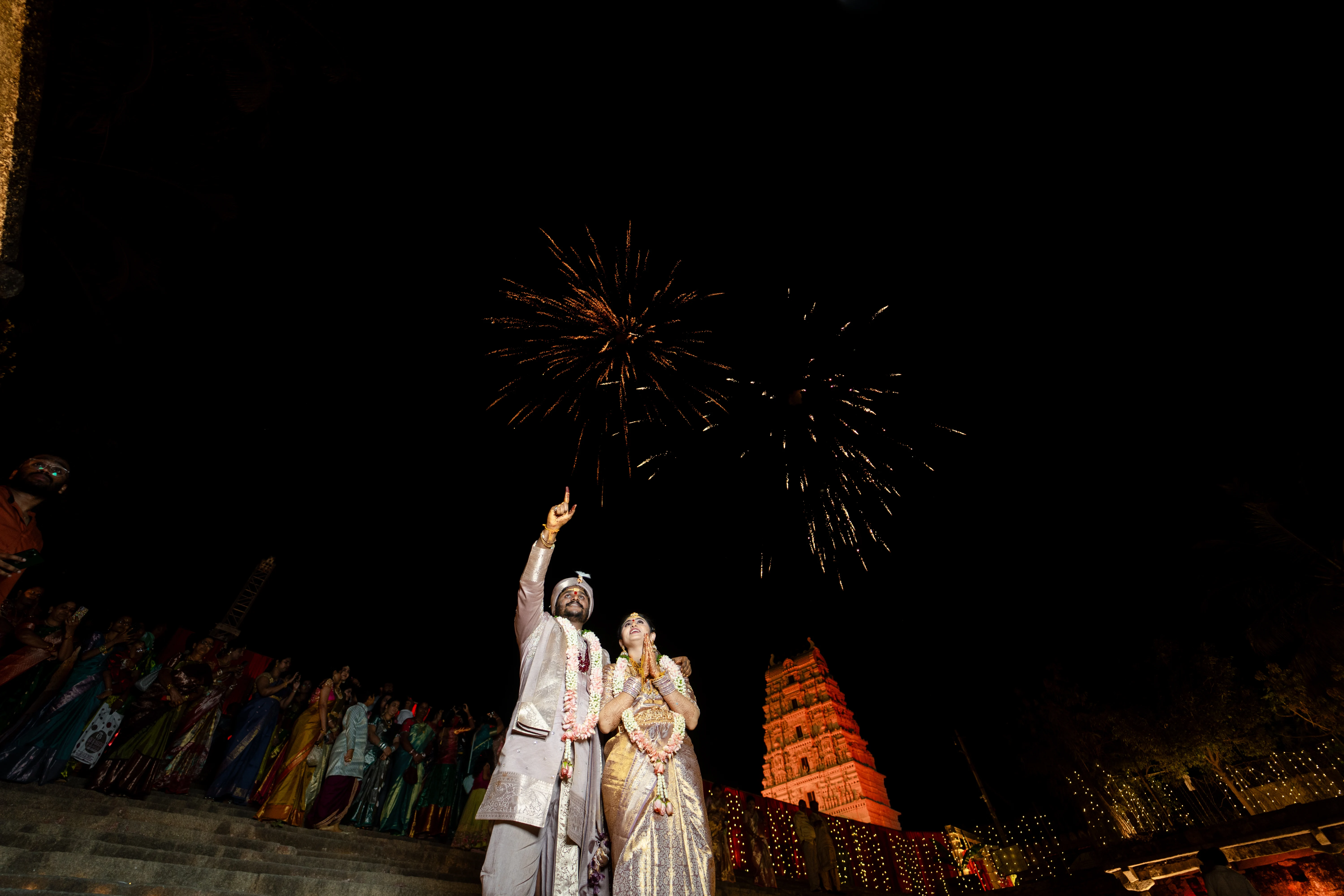 Couple embraces under a sky filled with fireworks, celebrating their union in front of an illuminated temple, surrounded by friends and family dressed in traditional attire, skillfully captured by Out of The Blues Fine Art Wedding Photography in Hyderabad.