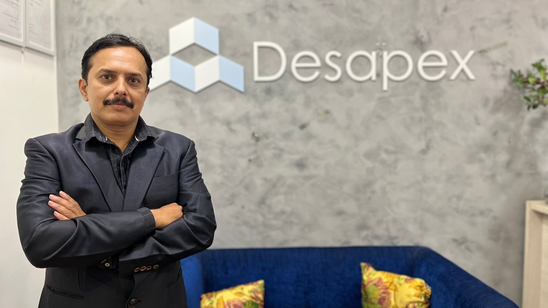 A man stands while two people sit at a table in an office setting, smiling and engaged in conversation. The mood is friendly and collaborative.