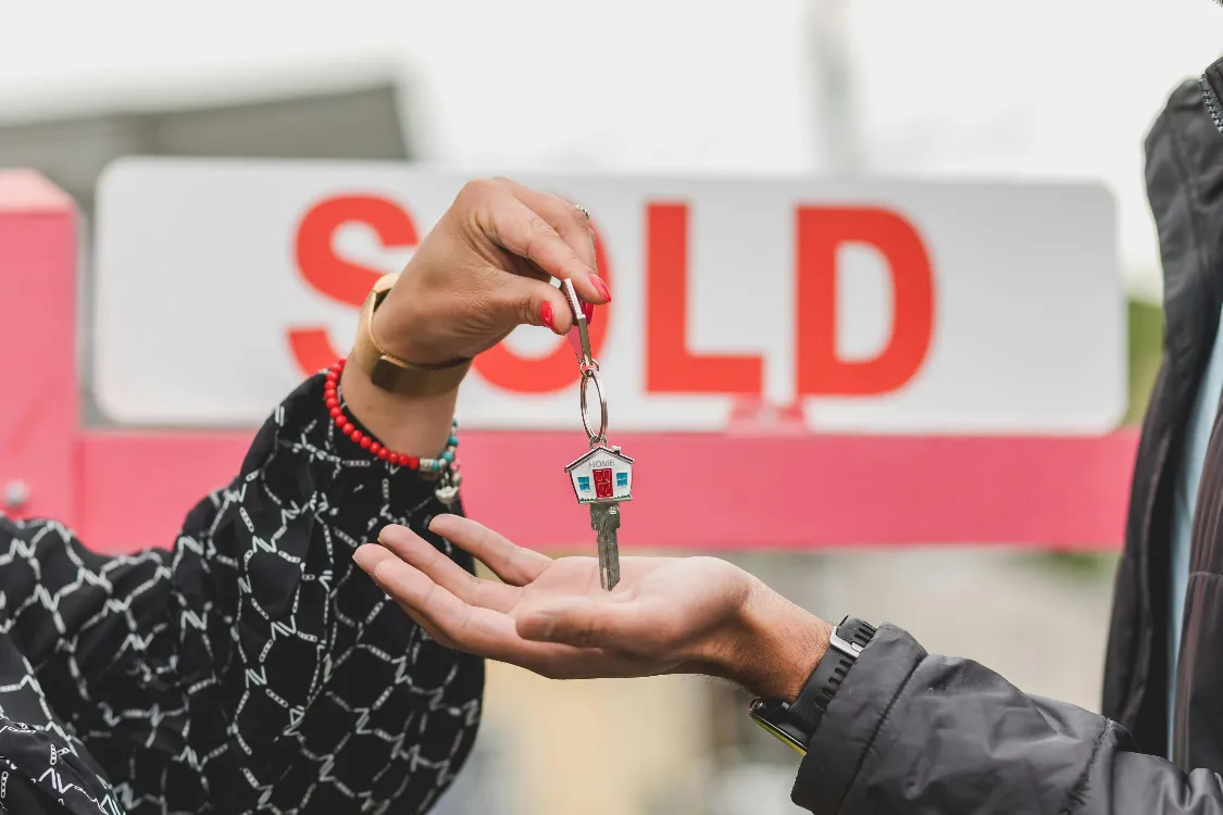 Close up of one person handing over house keys to another with a “SOLD” sign blurred in the background. This moment represents the final step of a property sale, often showcased on a real estate listing website to highlight successful transactions.