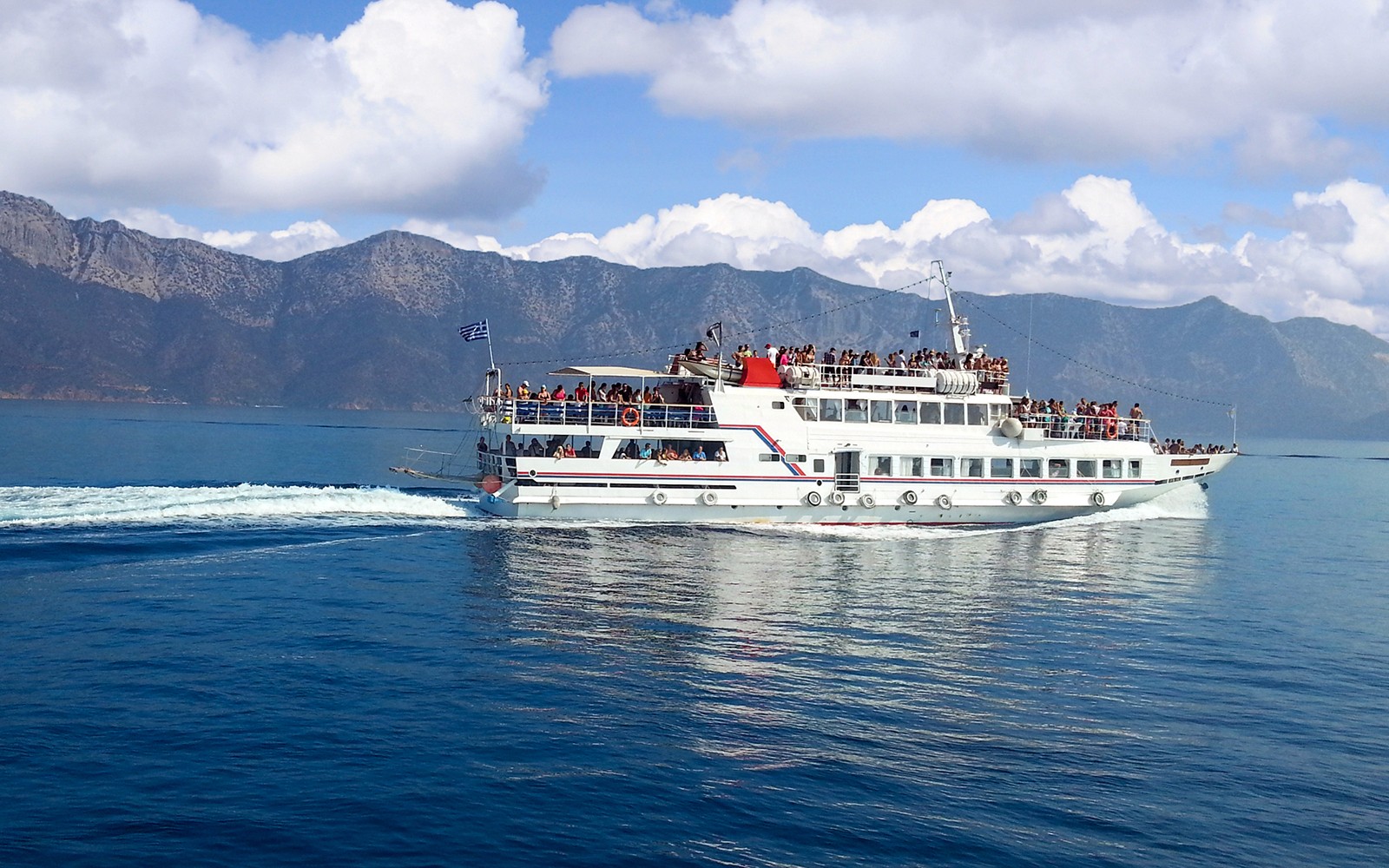 载有乘客的渡轮在希腊米科诺斯岛附近航行，背景是连绵的山峦。