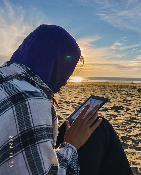 Rebecca sits on a beach as the sun is setting. She is wearing a hoodie and reading on her kindle.