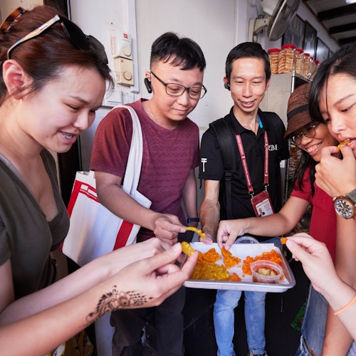 Four people share a tray of bright orange and yellow food. One person wears a hat and another has a bag. They appear to be enjoying the food together.