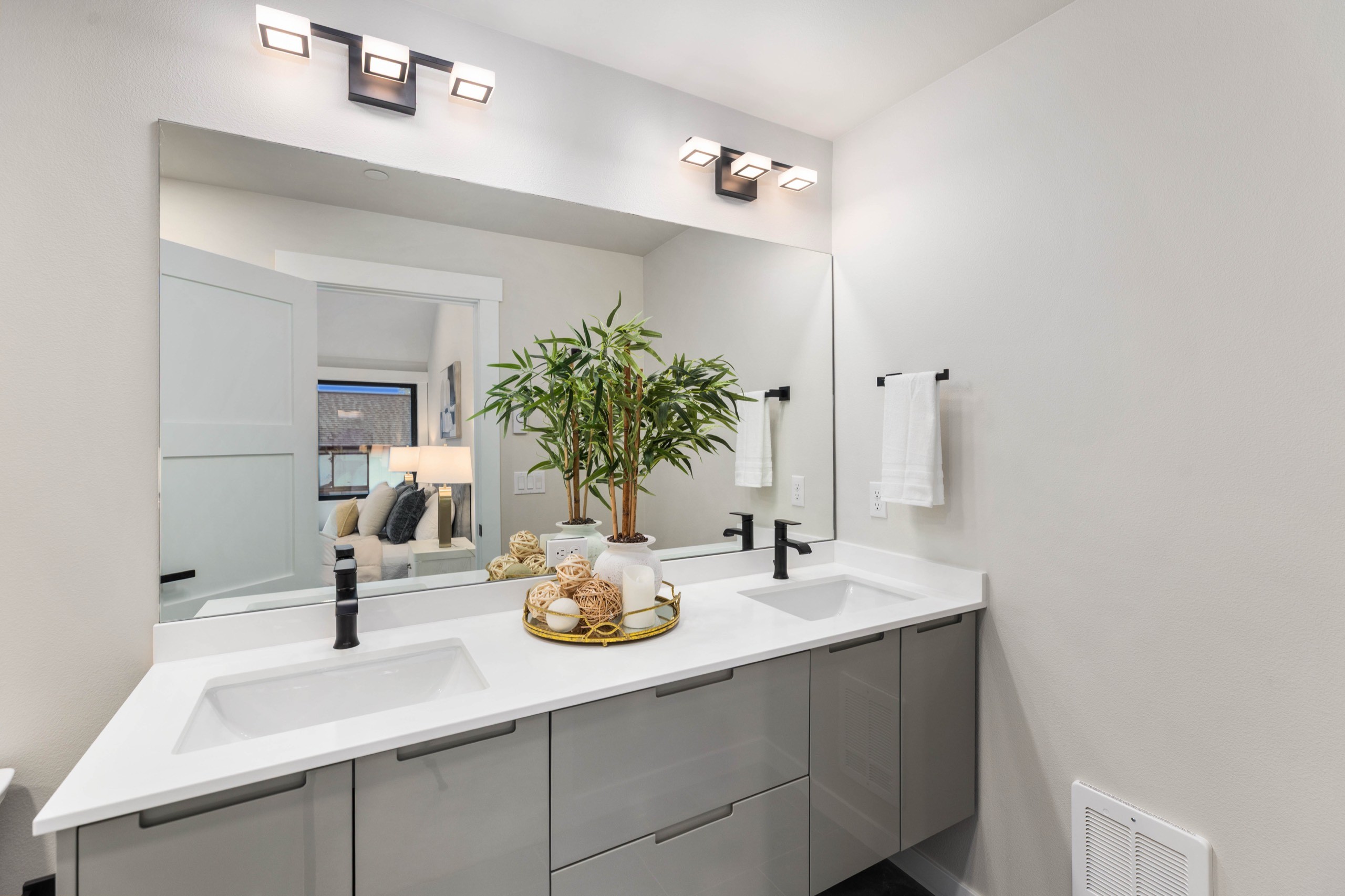 Luxury primary ensuite double vanity with grey cabinetry, white quartz countertop, dual black matte faucets, large backlit mirror, and decorative tray accent