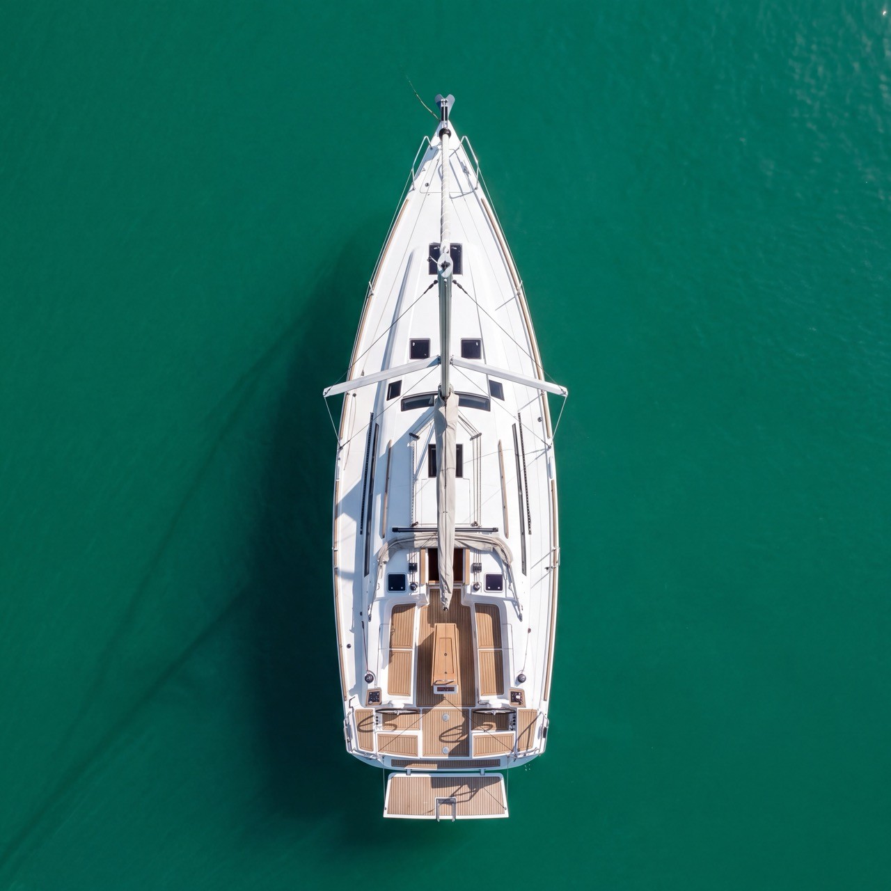 Aerial view looking straight down on a Dufour 350 GL sailing yacht anchored in emerald green water, showing its teak-covered cockpit sole, swim platform, and furled mainsail.