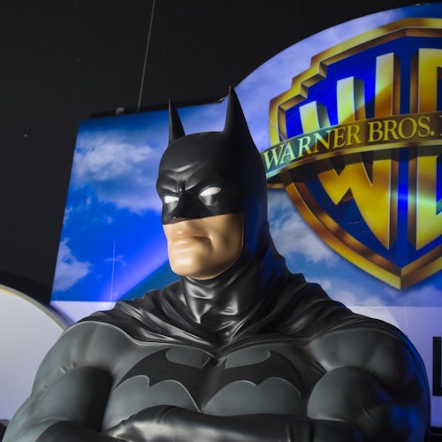 Statue von Batman mit verschränkten Armen vor einem Warner Bros.-Logo und einem Himmelshintergrund.