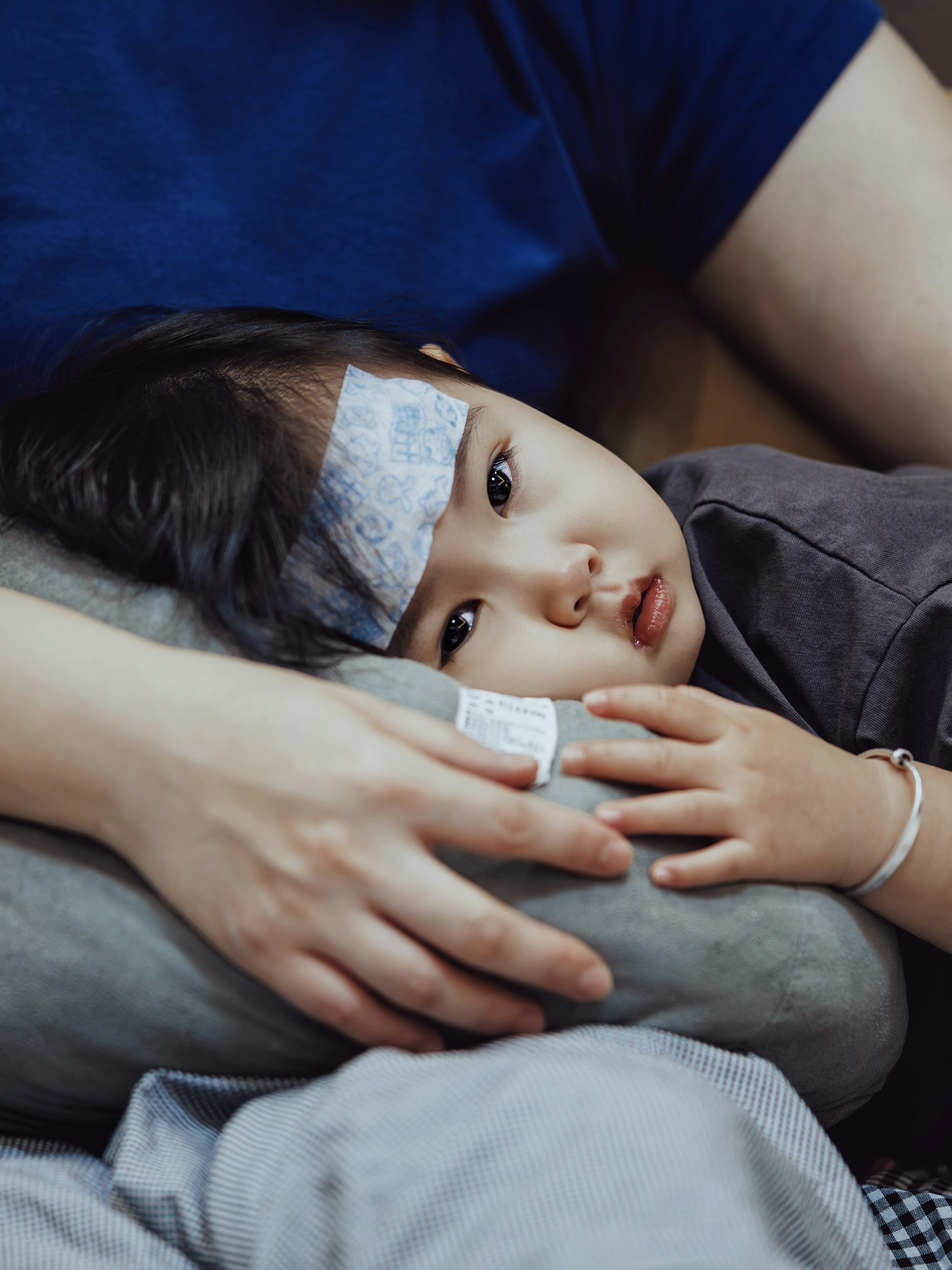 A young child with a cooling fever patch resting in a parent's arms at home while their fever is monitored