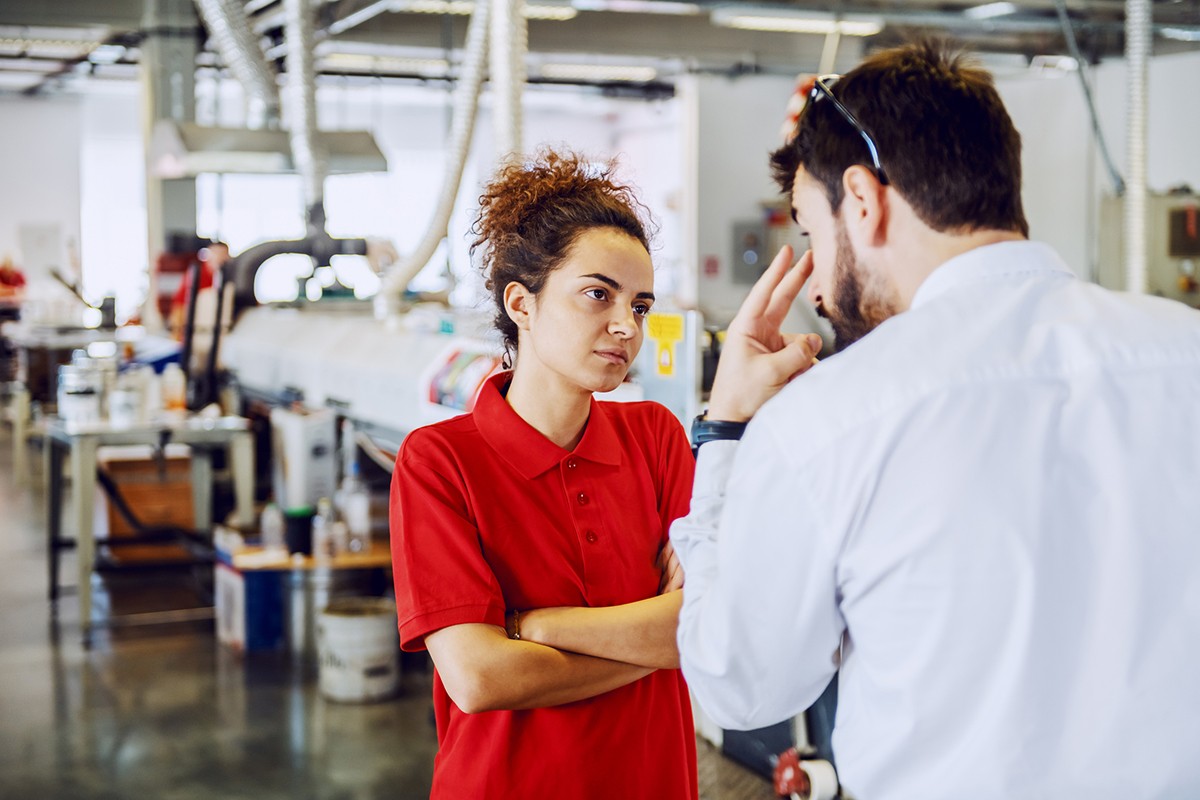 A woman with crossed arms looks warily at a male colleague gesturing towards her in a factory setting, suggesting a tense workplace confrontation.