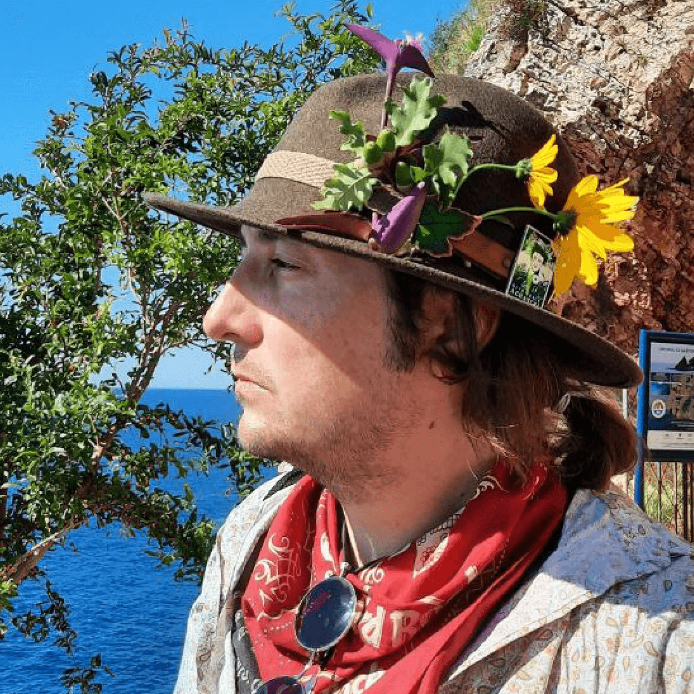 Konrad stairs into the distance facing profile with flowers pinned to hs wide brimmed hat and a red bandanna around his neck..