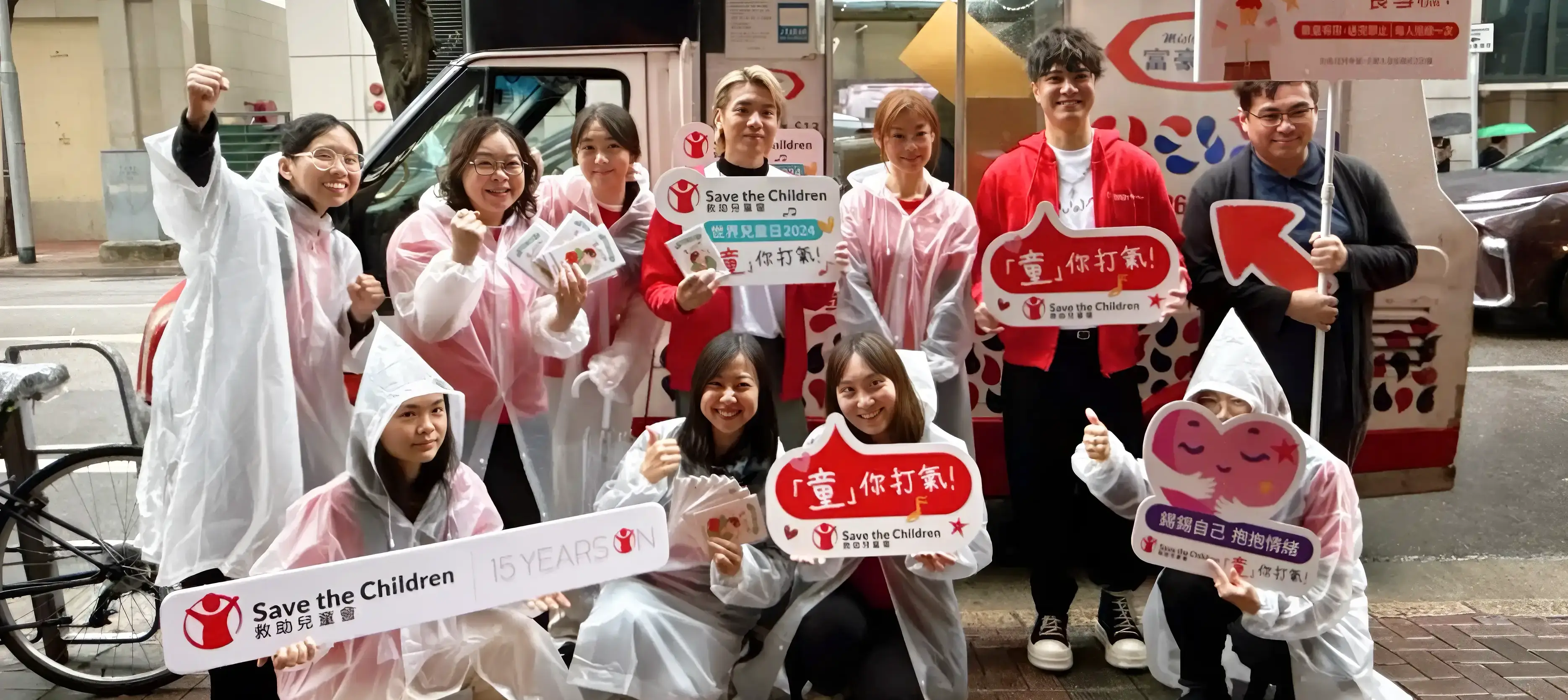 Save the Children Hong Kong World Children’s Day 2024 campaign hero – group of volunteers in raincoats holding “Cheer You Up” and “童你打氣” photo props, promoting student mental health awareness by Teatime Studio.