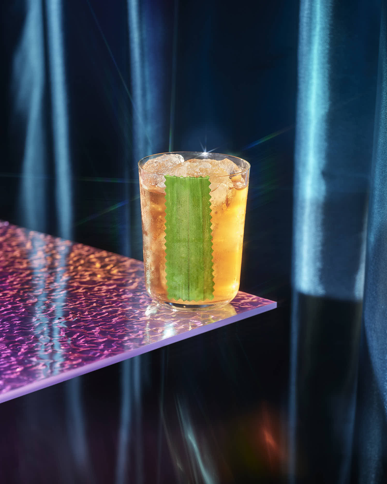 A vibrant cocktail sits on a reflective surface, illuminated by colorful lights in a dark setting.