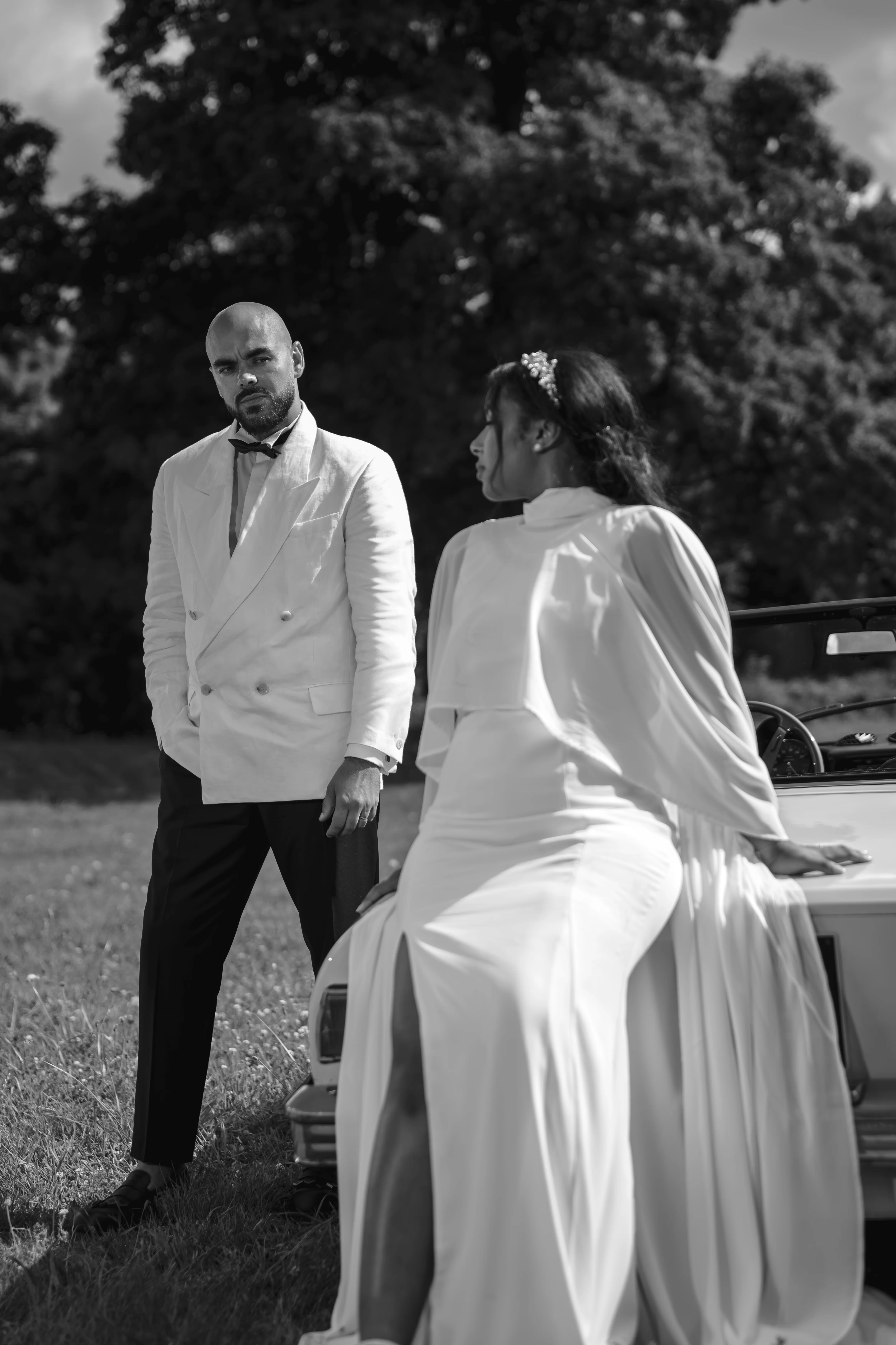 Black and white photo of a man in a white double-breasted jacket standing beside a woman in a white gown seated on a vintage car
