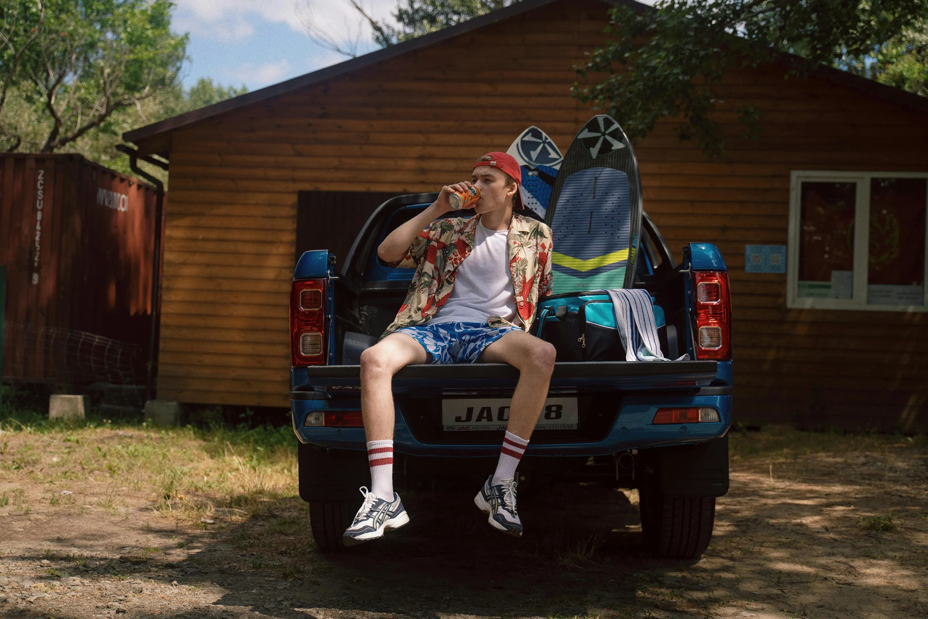 A young man in a Hawaiian shirt drinks while sitting in the back of a pickup truck with surfboards