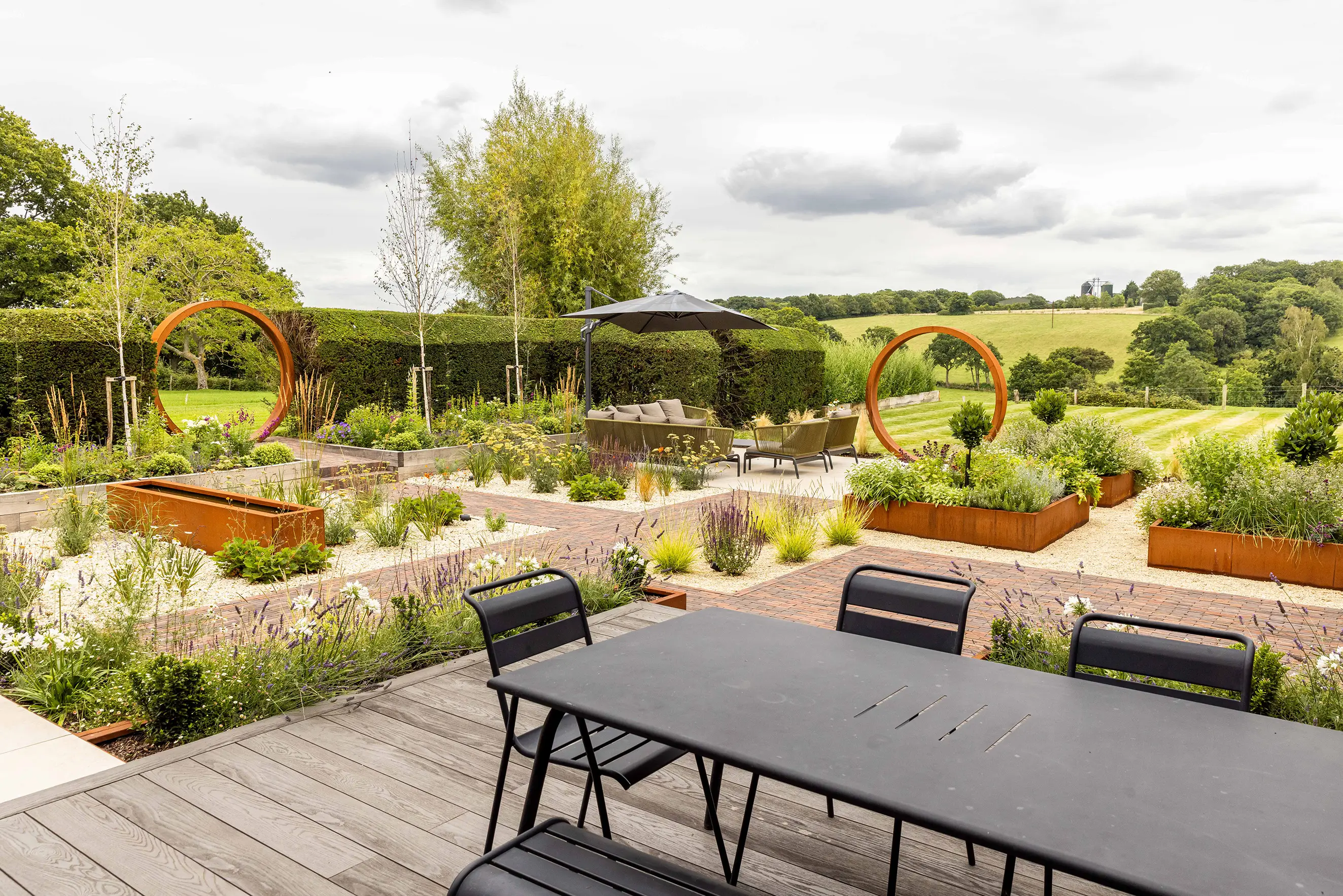 A serene garden view featuring lush greenery, flower beds, and a dining table set against a cloudy sky.