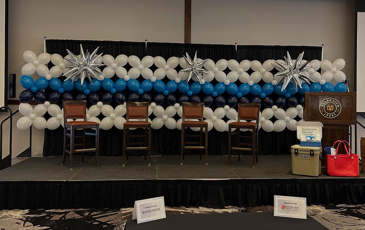 A stage setup for a corporate event features a backdrop of white, blue, and silver star-shaped balloons, with four wooden chairs facing the audience, a podium on the right, and a table displaying informational brochures and a red bag in the foreground.