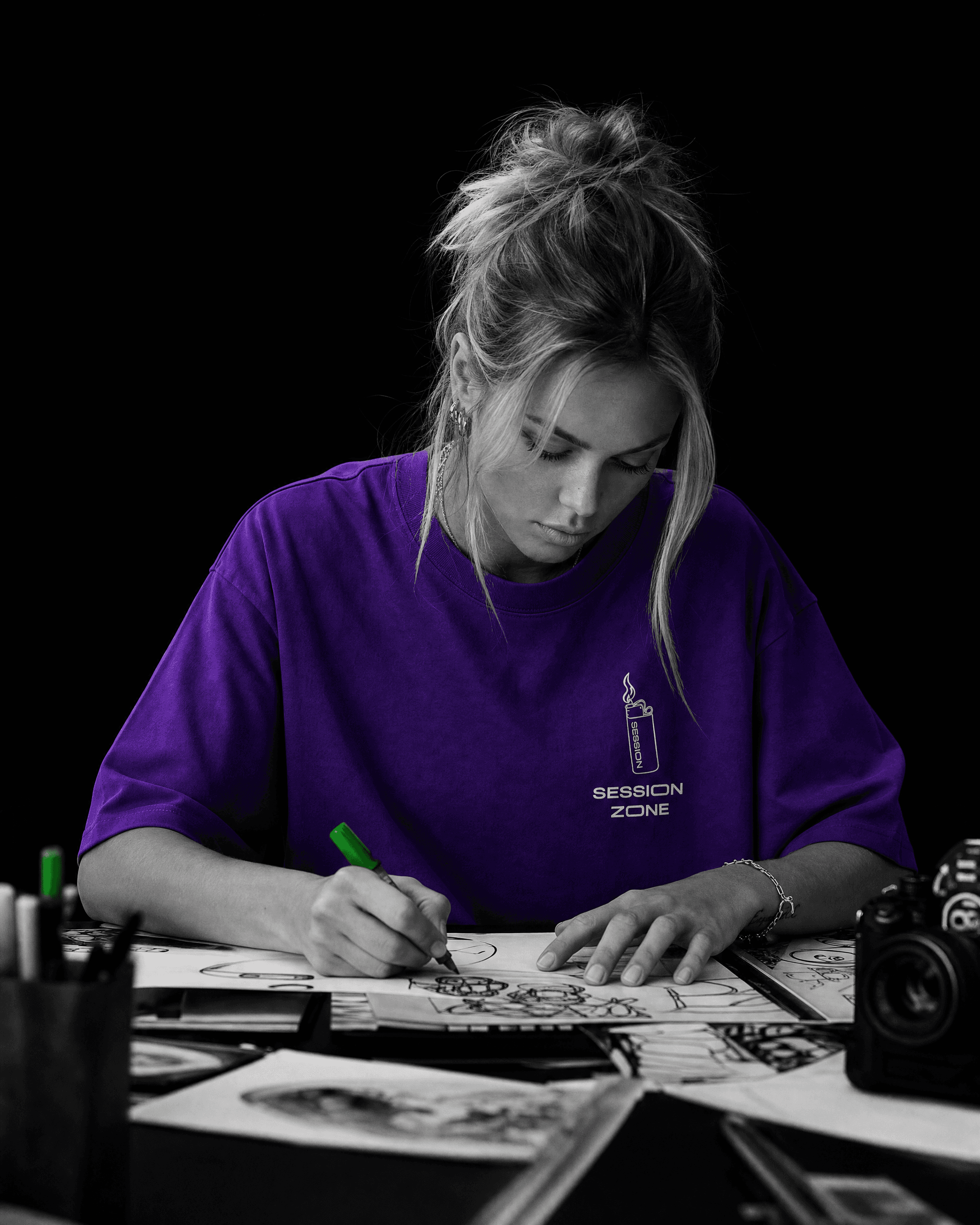 Woman wearing a purple Session Zone lighter-graphic T-shirt while sketching beside a camera and photographic prints.