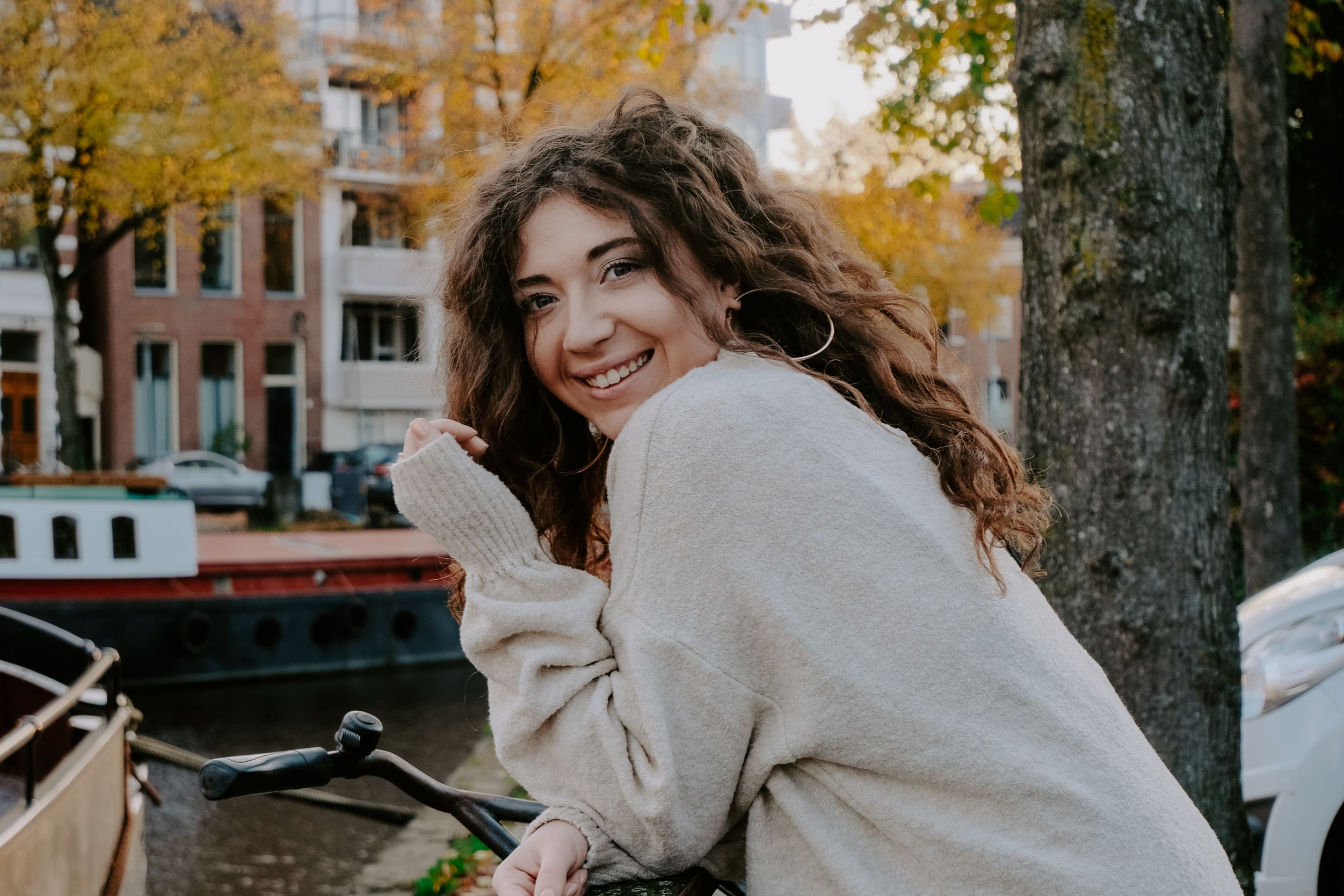 Een jonge vrouw met golvend haar glimlacht terwijl ze bij een gracht zit met herfstbladeren op de achtergrond.