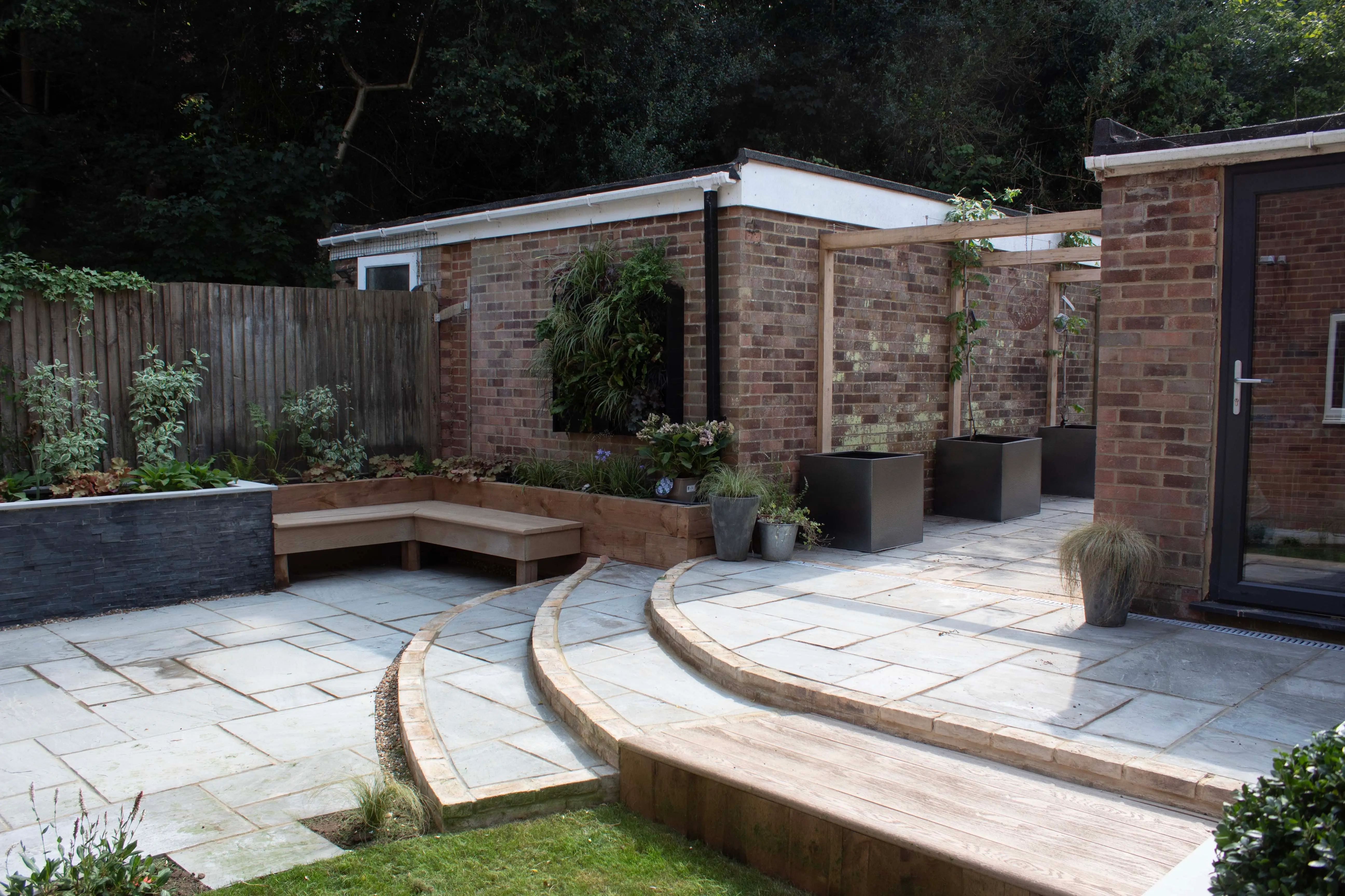 A modern garden with stone pathways, a wooden deck, and lush greenery surrounding a brick structure.