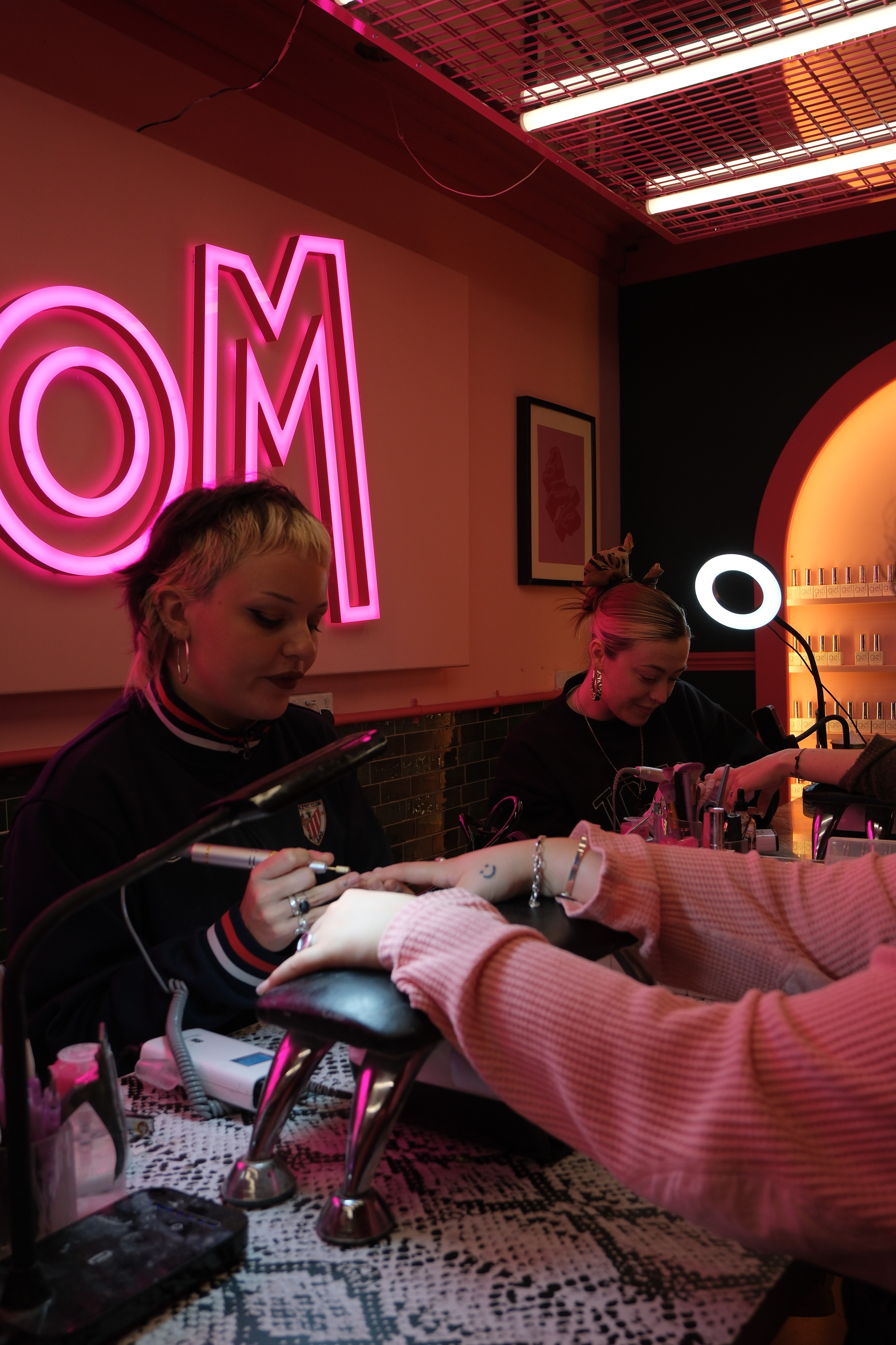 A wide, atmospheric shot of the salon during a busy session. Two nail technicians are at work with their clients at a snakeskin-patterned desk. The scene is dominated by a large, glowing pink neon "VENOM" sign, with warm desk lamps providing task lighting in the moody, dark-toned room.