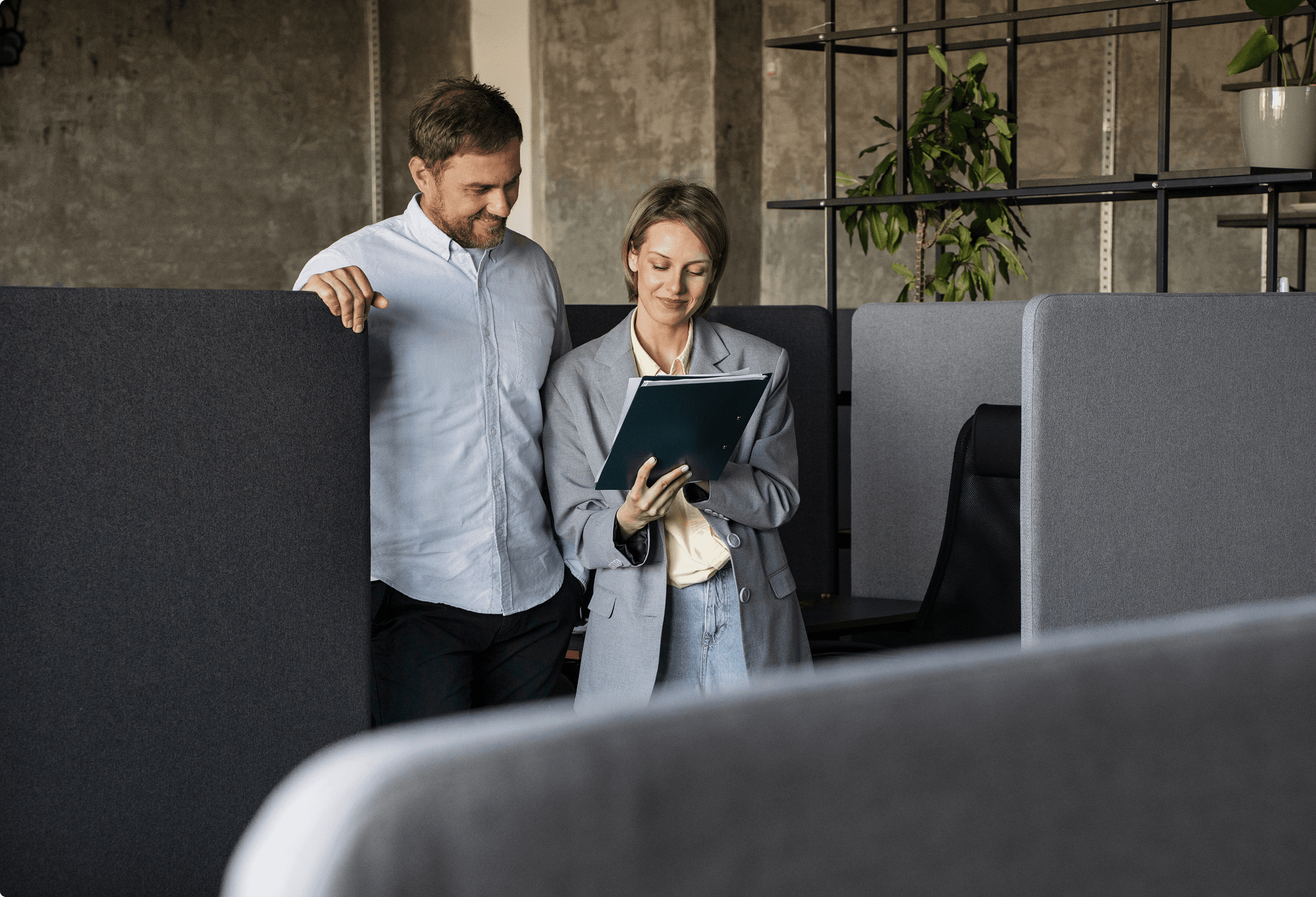 Two colleagues discuss work by a cubicle in a modern office setting. One holds a tablet, the other leans in attentively.